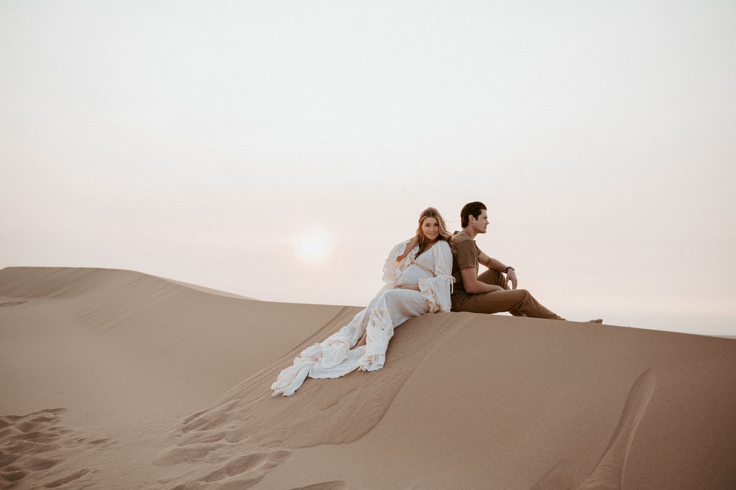 A romantic desert photoshoot features a couple sitting on sandy dunes at dusk.