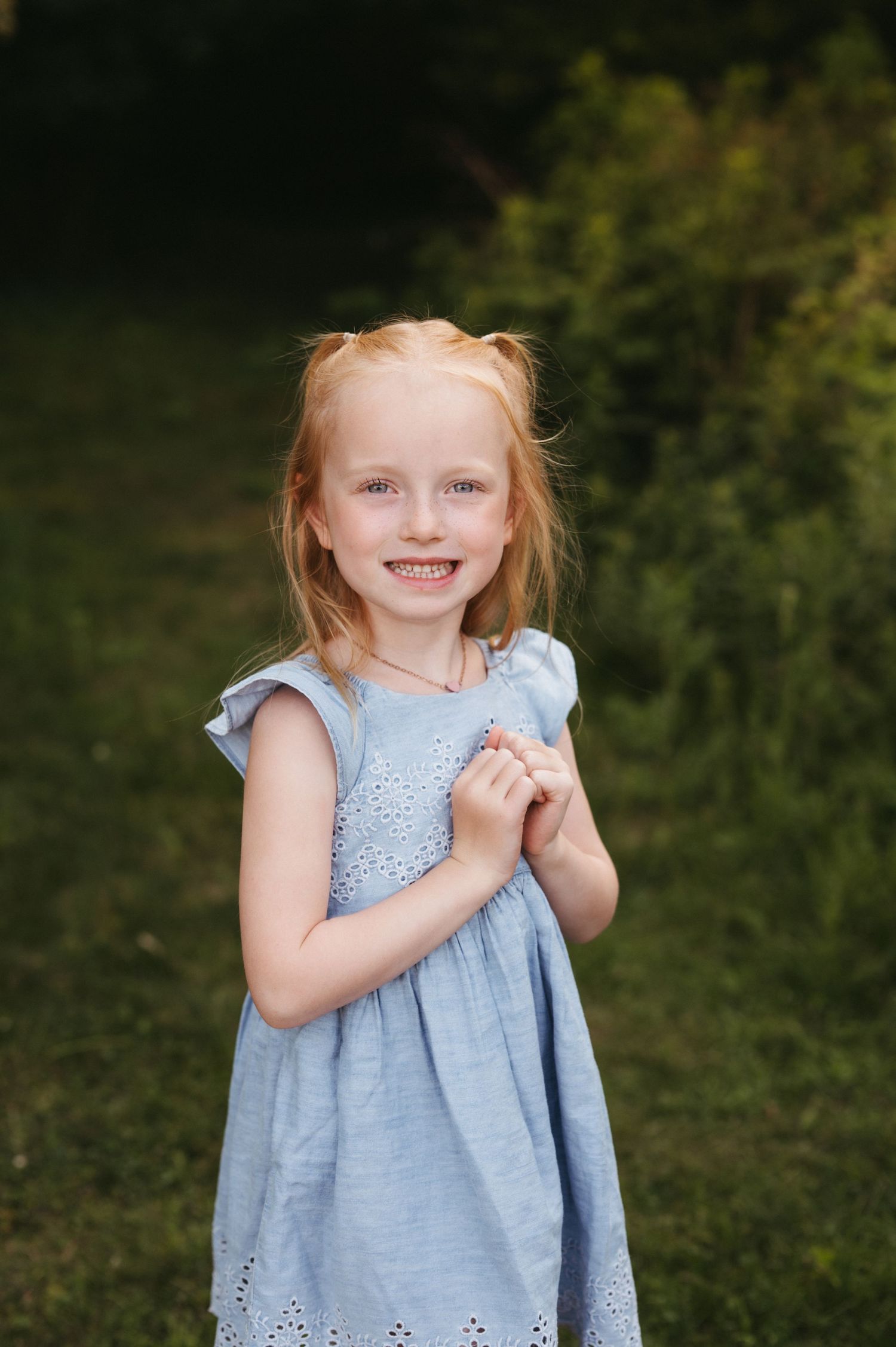 Young child in a light blue eyelet dress smiles during outdoor photo session.