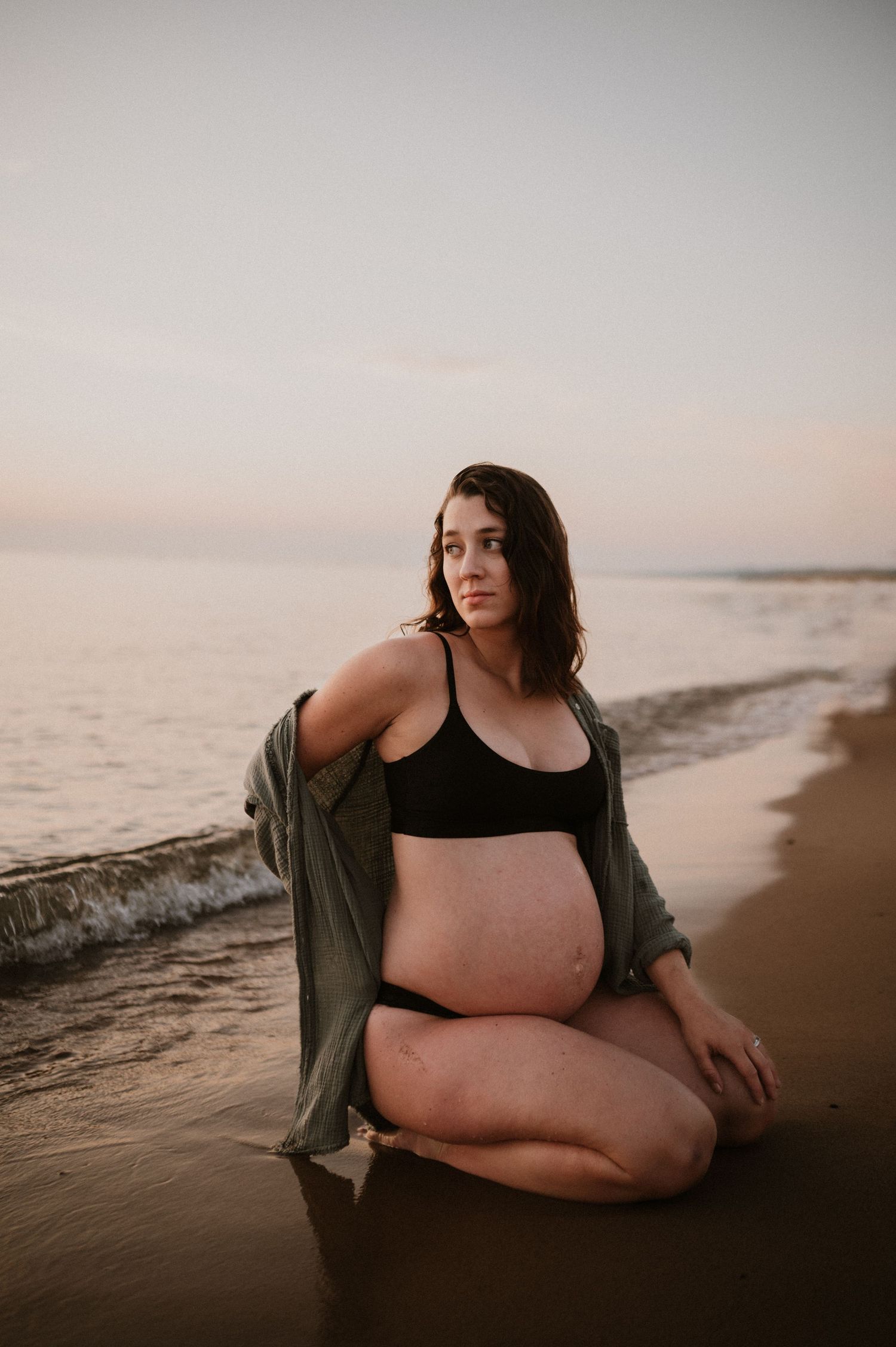 Maternity beach portrait with pregnant woman wearing olive cardigan sitting by ocean.
