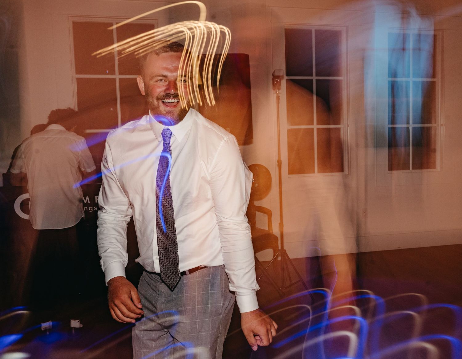 Long exposure photo creates light trails around dancing wedding guest in white shirt and blue tie.