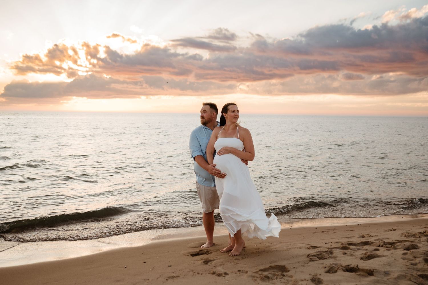 A couple embraces on a sandy beach at sunset with dramatic clouds and ocean waves in the background.
