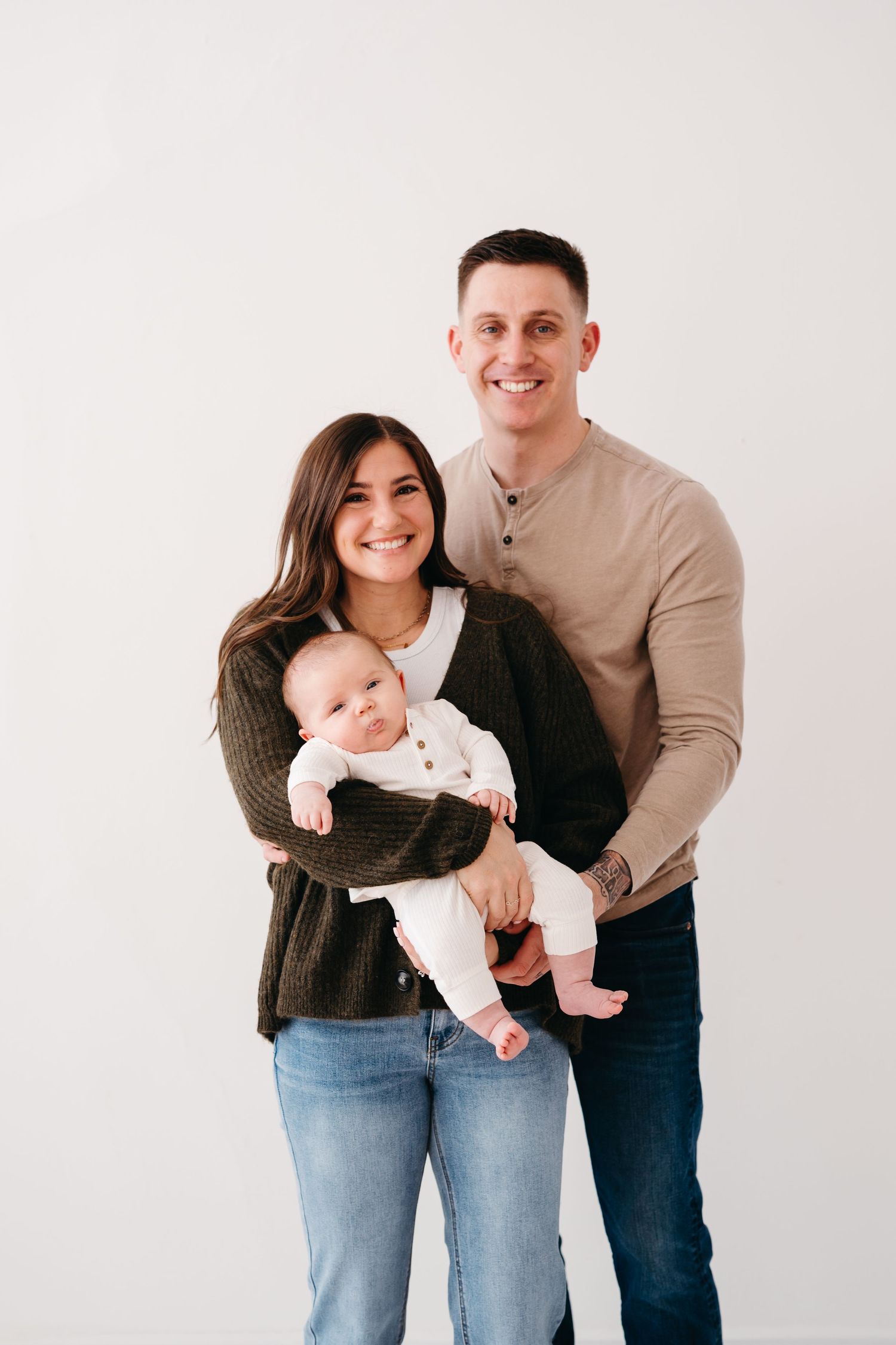 A young family poses together against a white backdrop, with the parent holding their baby in a white onesie.