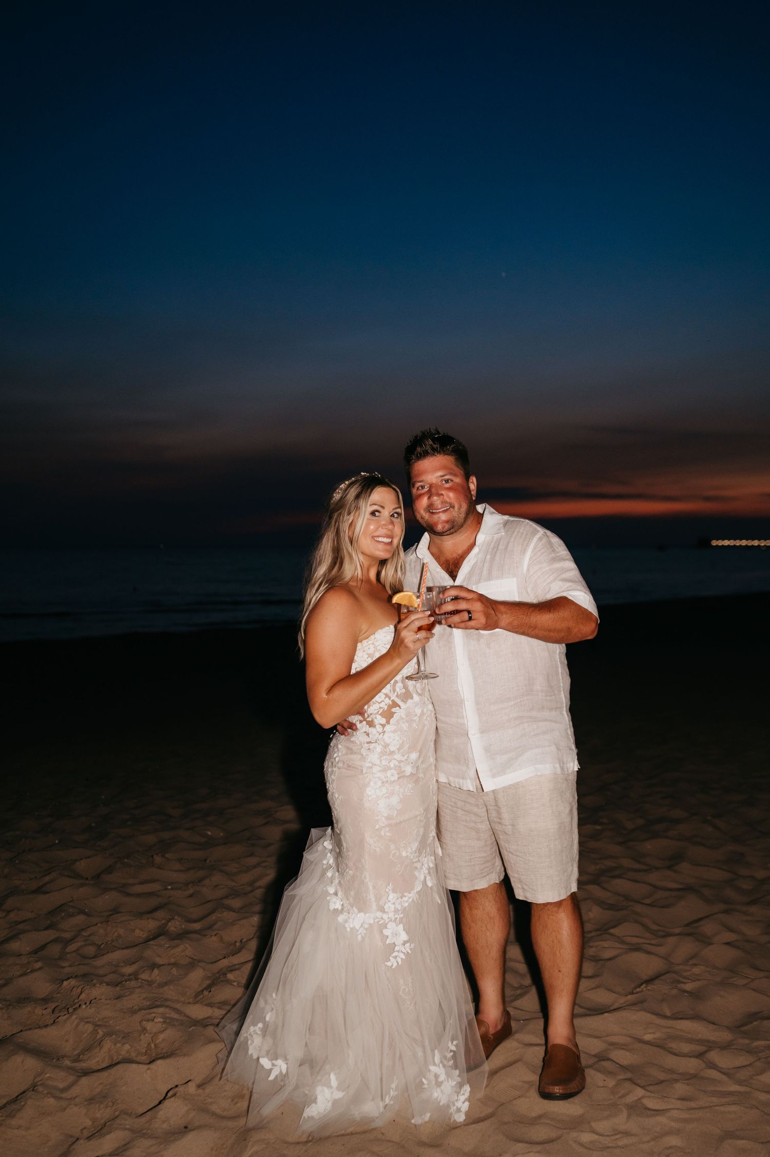 A couple enjoys drinks together while sitting on a sandy beach at dusk.