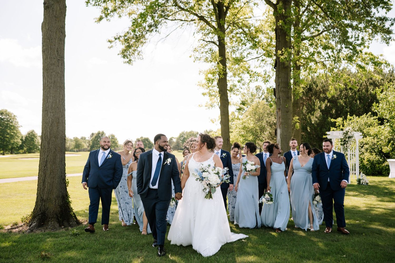 Wedding party gathered for group photos under tall trees on a golf course.