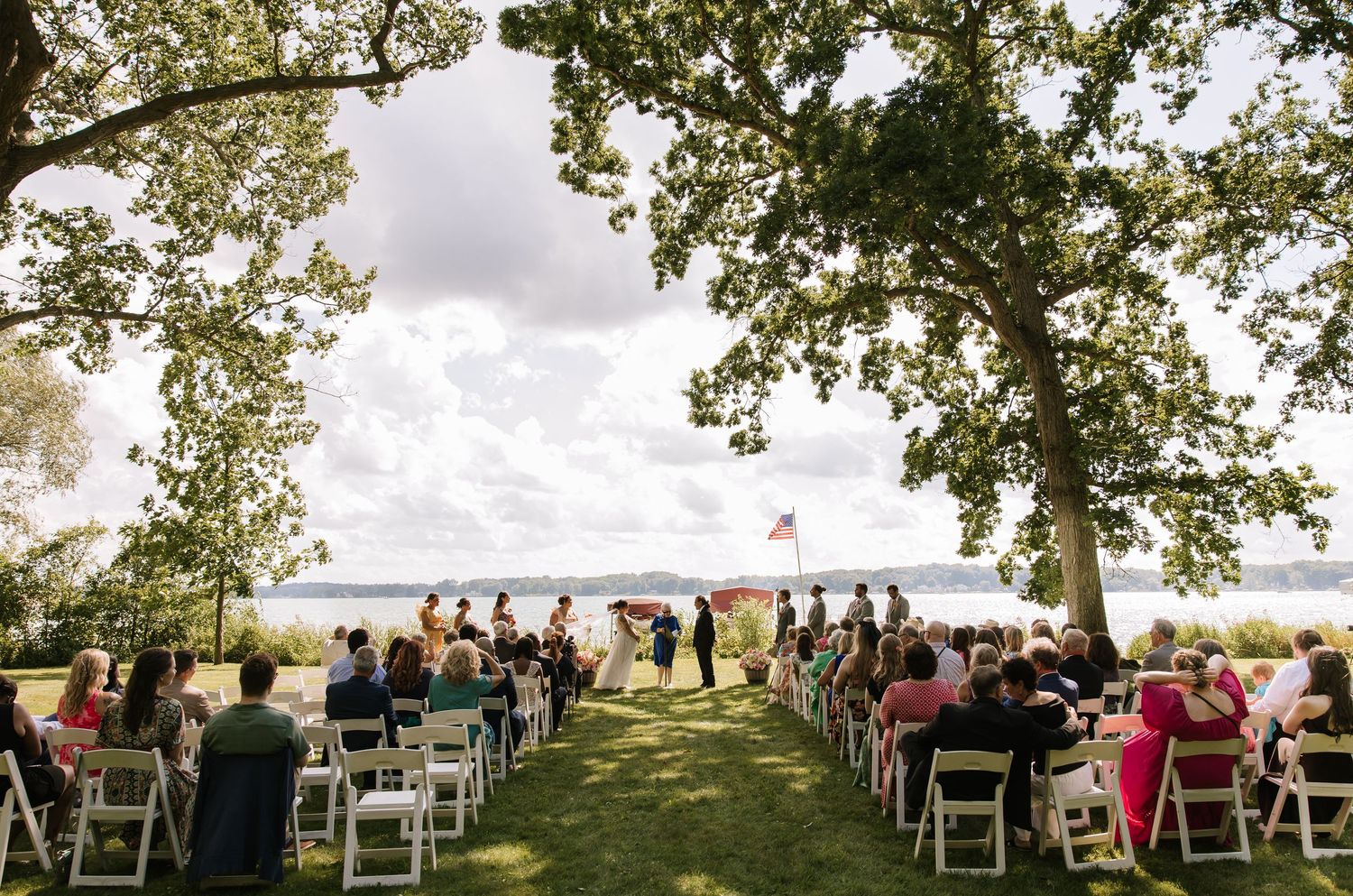 A panoramic view of an outdoor wedding ceremony setup with white chairs arranged under tall trees beside a waterfront.