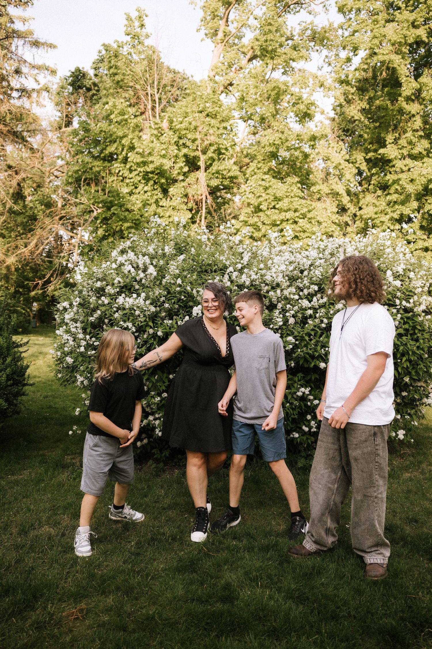 A family of four laughs together outdoors near blooming white bushes and lush green trees on a sunny day.