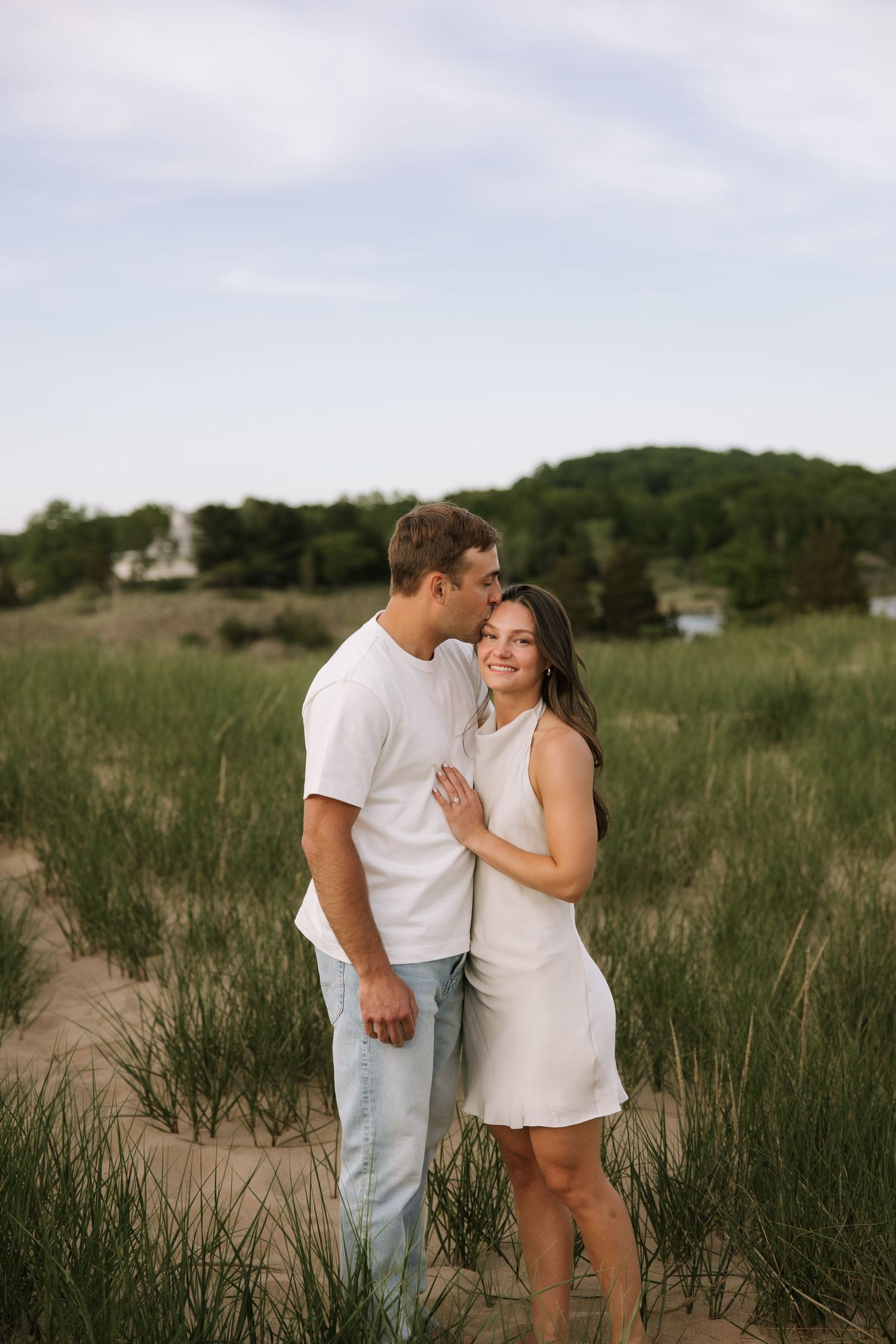 Romantic couple sharing a kiss on sandy beach dunes, man in white tee and jeans, woman in white dress.