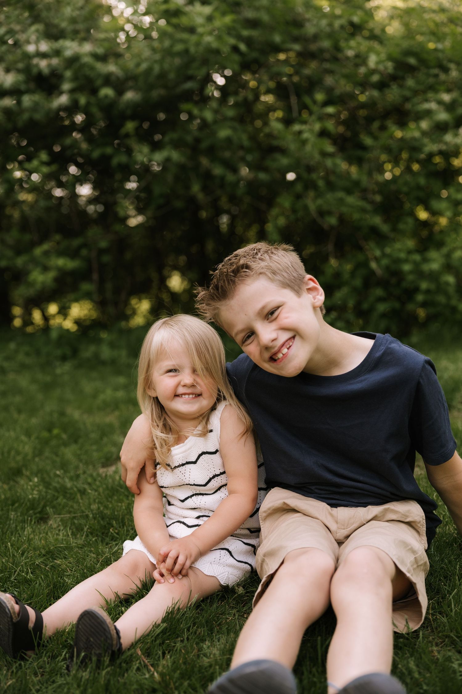 Two smiling children, a young girl and older boy, sit together on green grass with lush trees in the background.