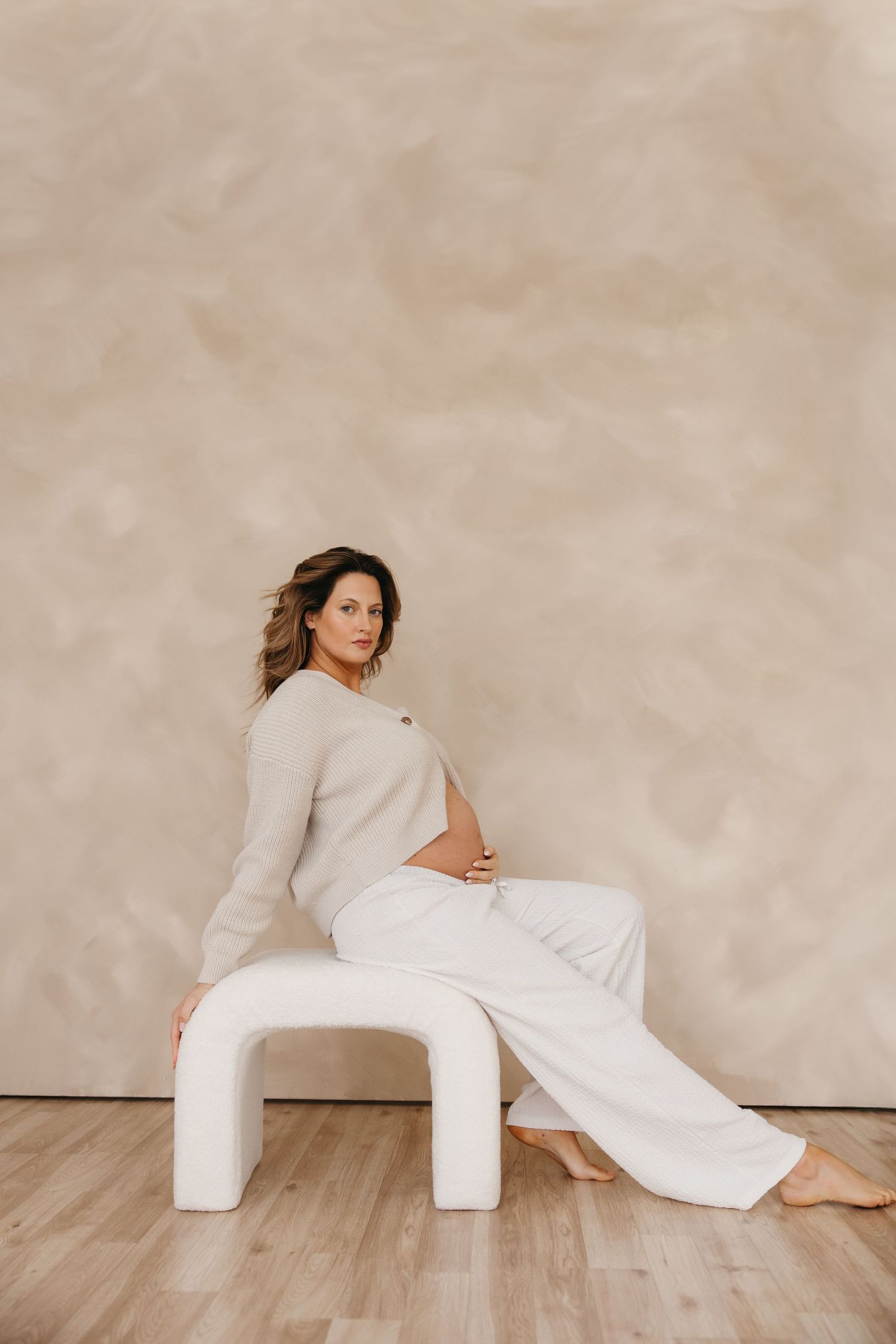 Pregnant woman in beige sweater and white wide-leg pants sitting on a white arch stool, side profile.