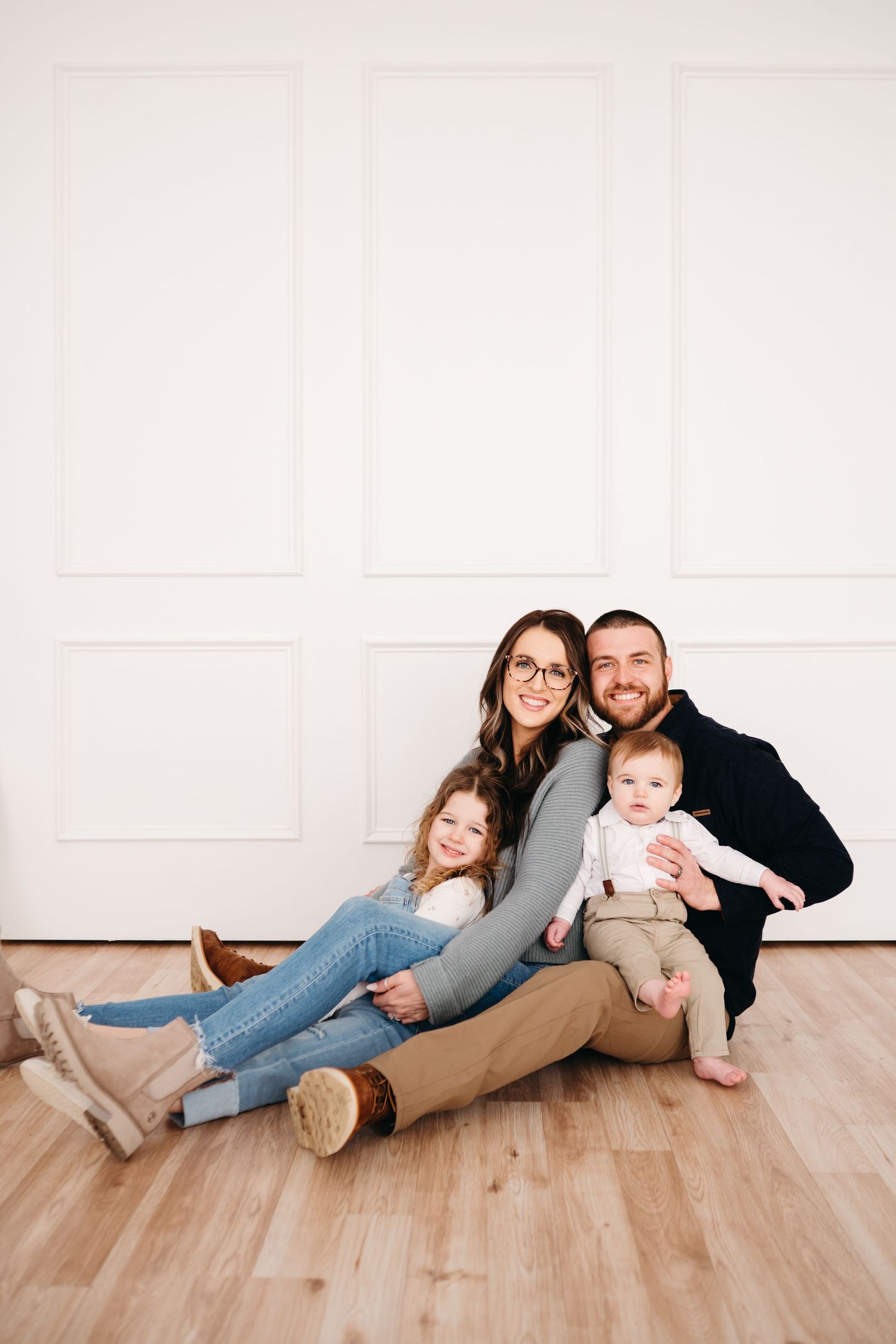 A family sits together on a hardwood floor against a white wall, casually posed in coordinating outfits.