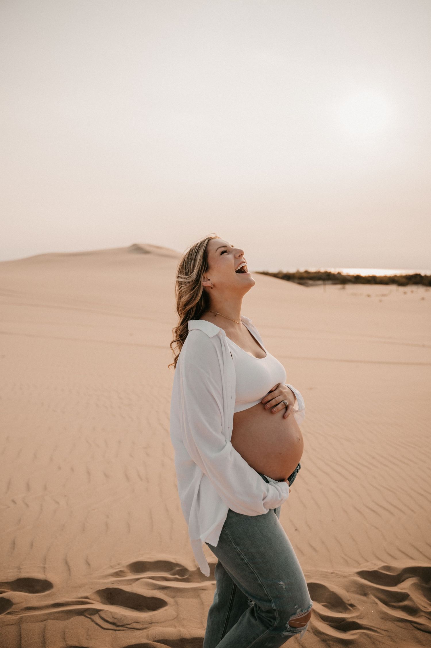 Pregnant woman in white shirt and jeans stands joyfully against desert backdrop.