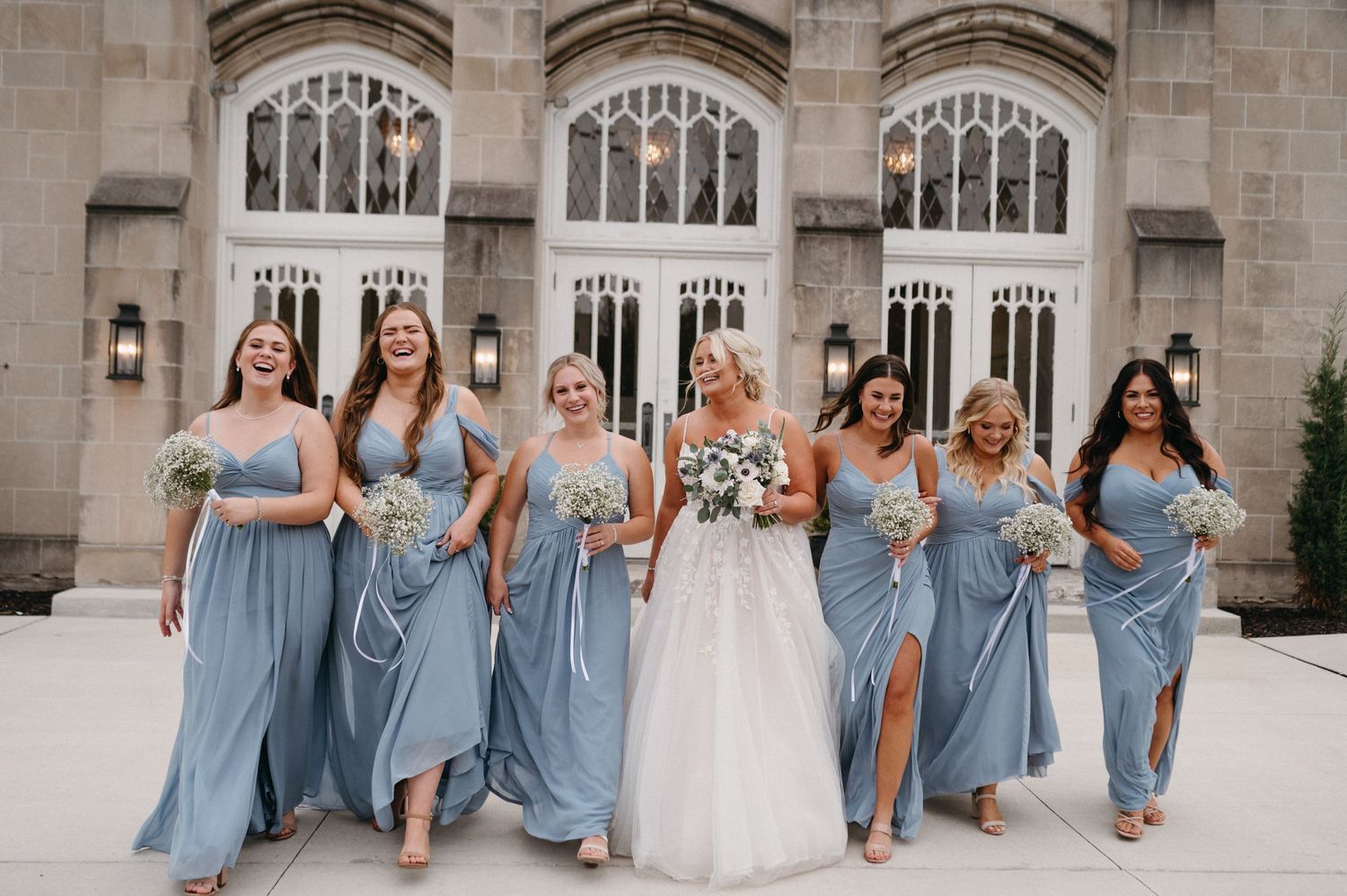 Wedding party in light blue dresses walking and smiling against Gothic church backdrop.