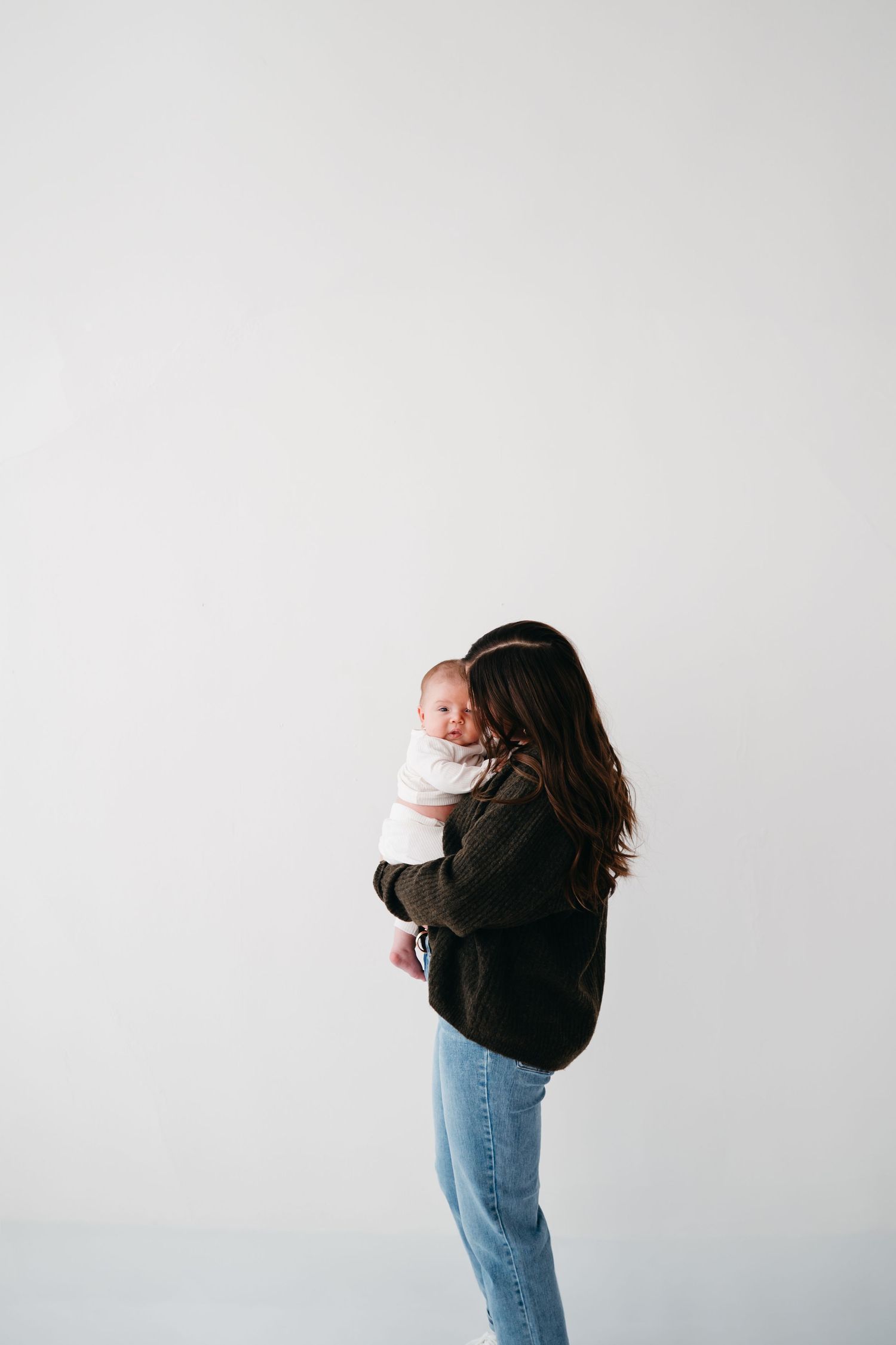 A person wearing a black sweater and jeans holds a baby against a white background.