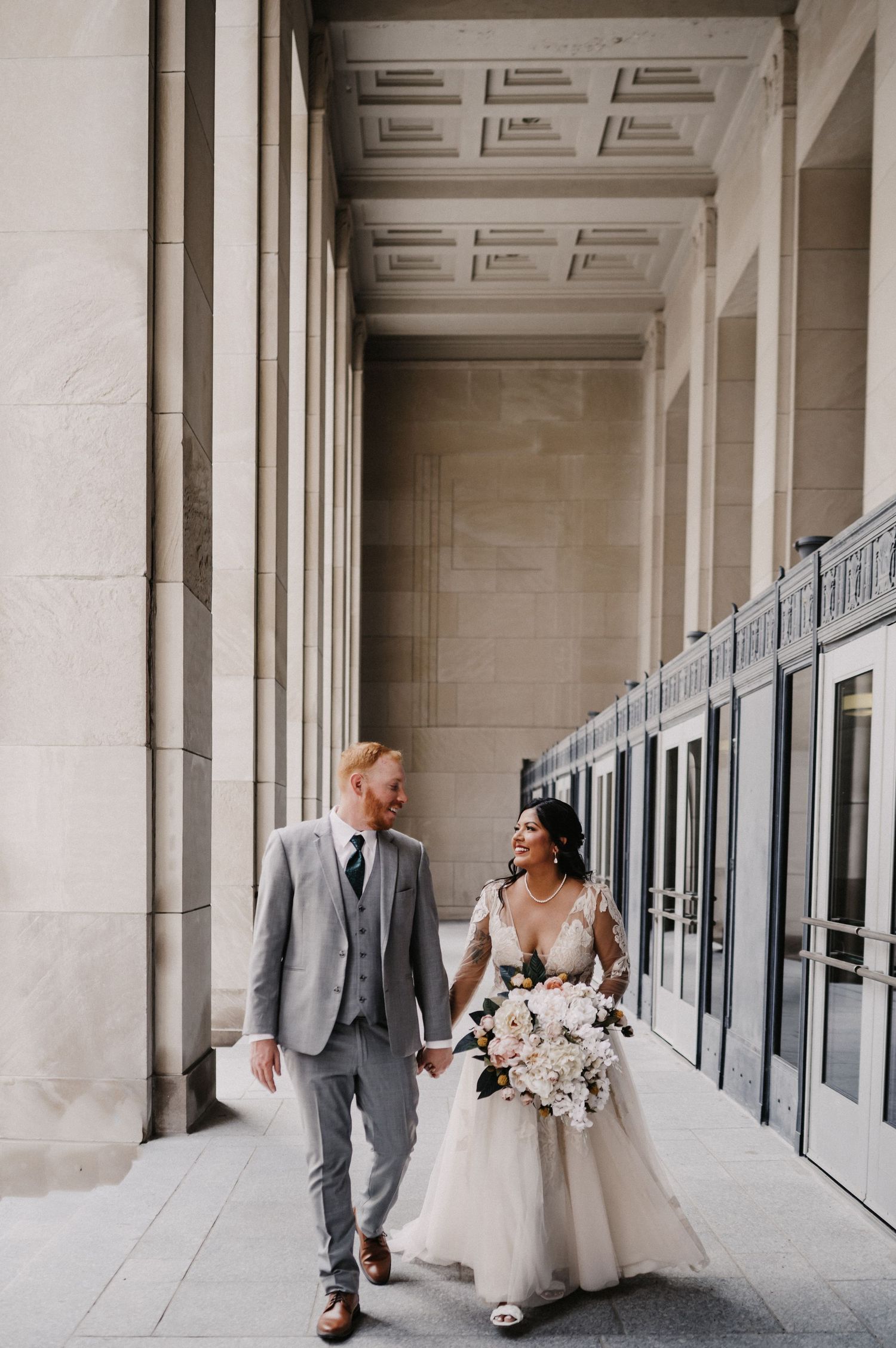Newlyweds walk hand in hand down elegant hallway with classical architecture.