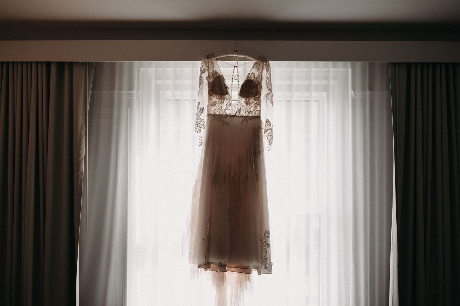 Delicate lace wedding dress hanging in front of white curtained window.