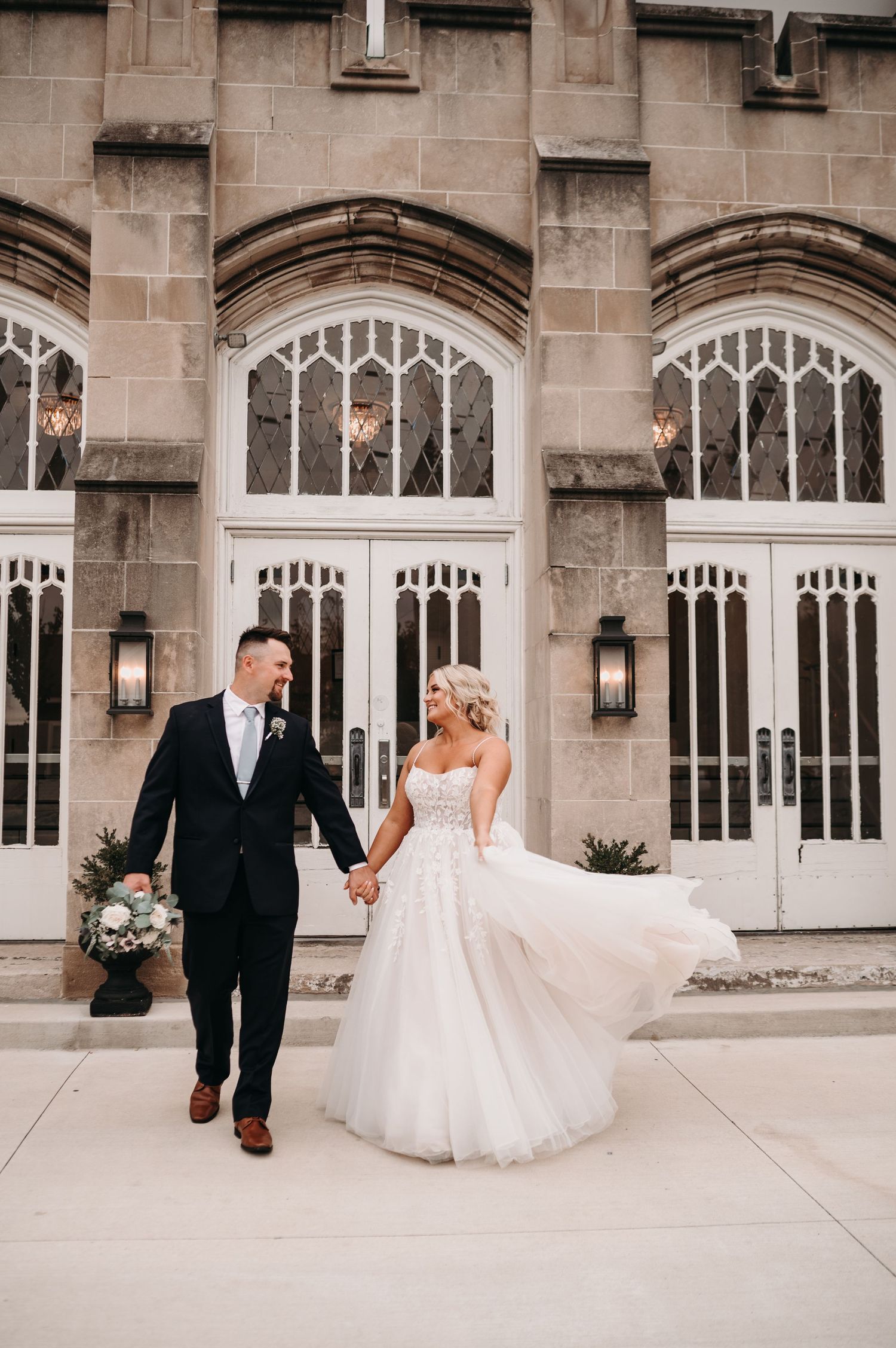 A couple walks hand in hand outside a beautiful stone building with Gothic architecture.