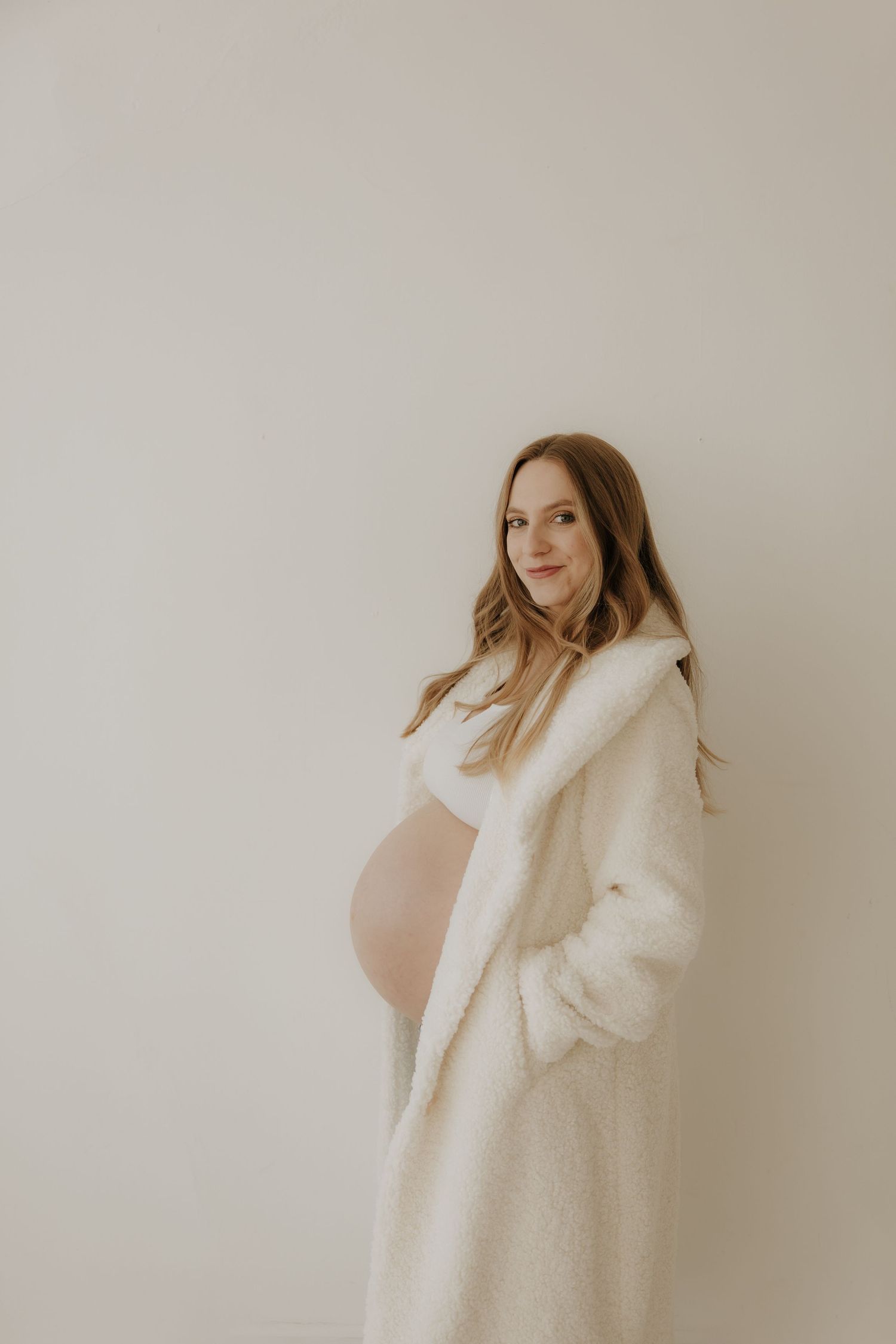 Expectant mother wearing cozy white sweater poses against minimalist backdrop.