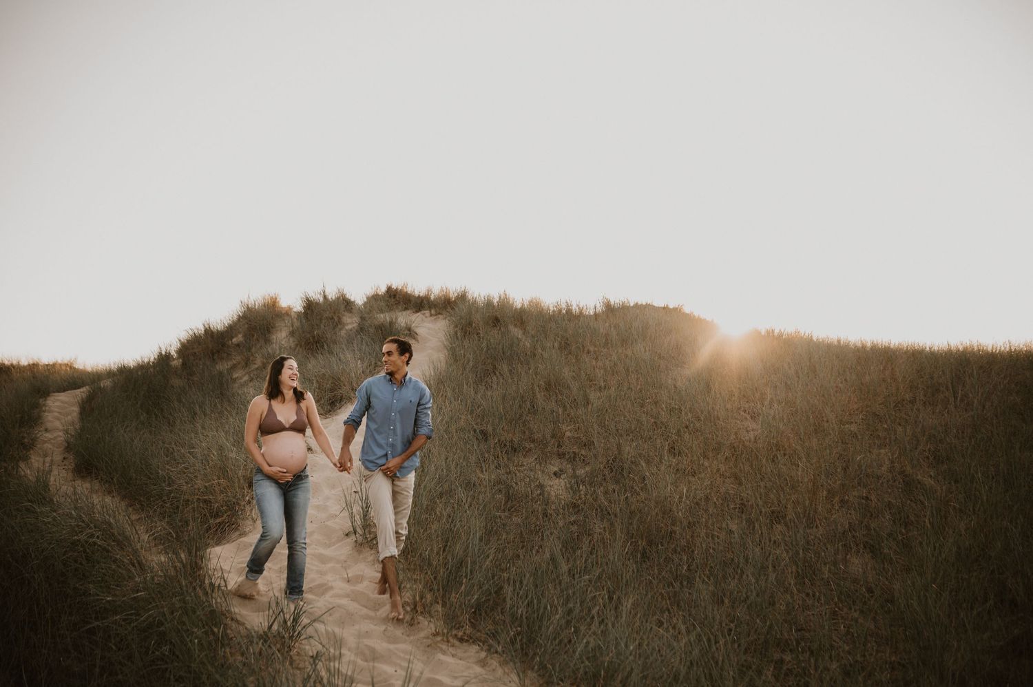 Couple walking along grassy coastal path during sunset maternity photoshoot.