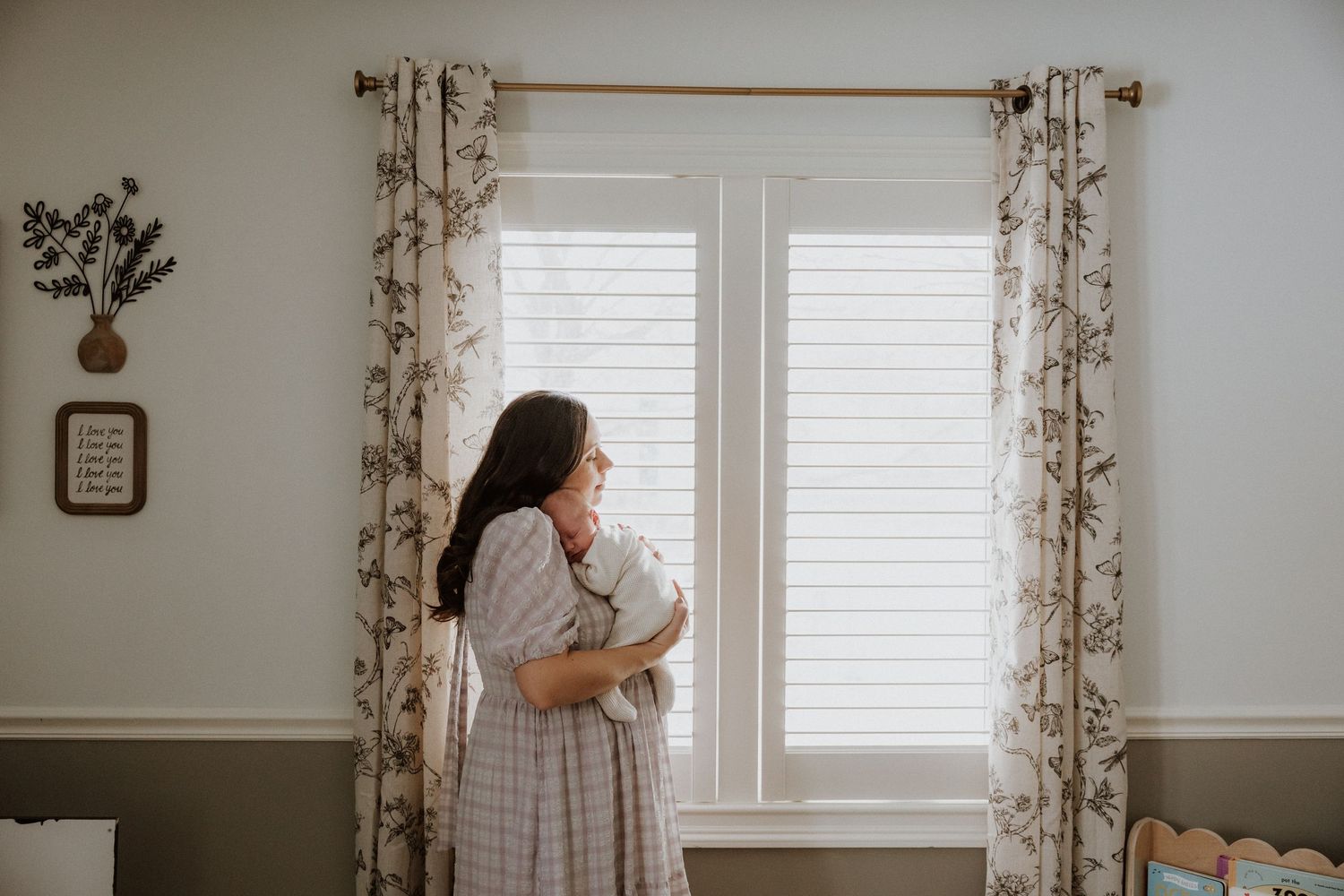 A tender moment by a window with floral curtains as a newborn is held in a bright, airy nursery.