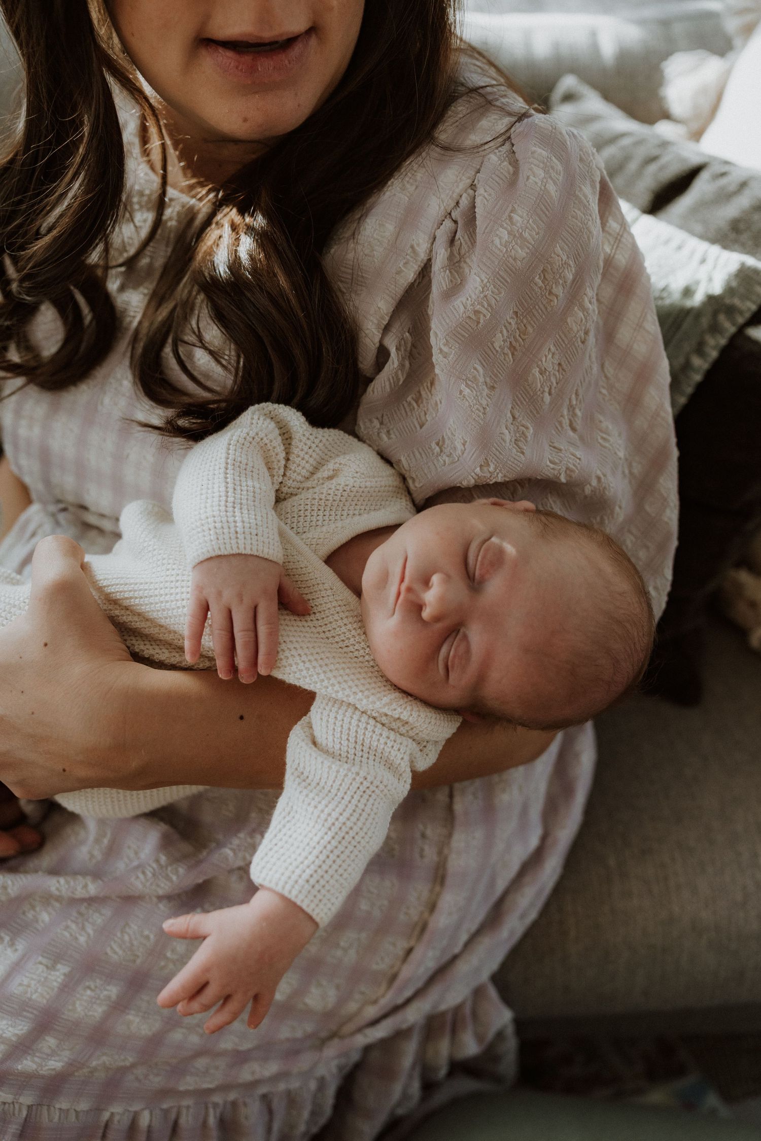 An intimate moment showing a sleeping baby in a white knit outfit on a grey couch.