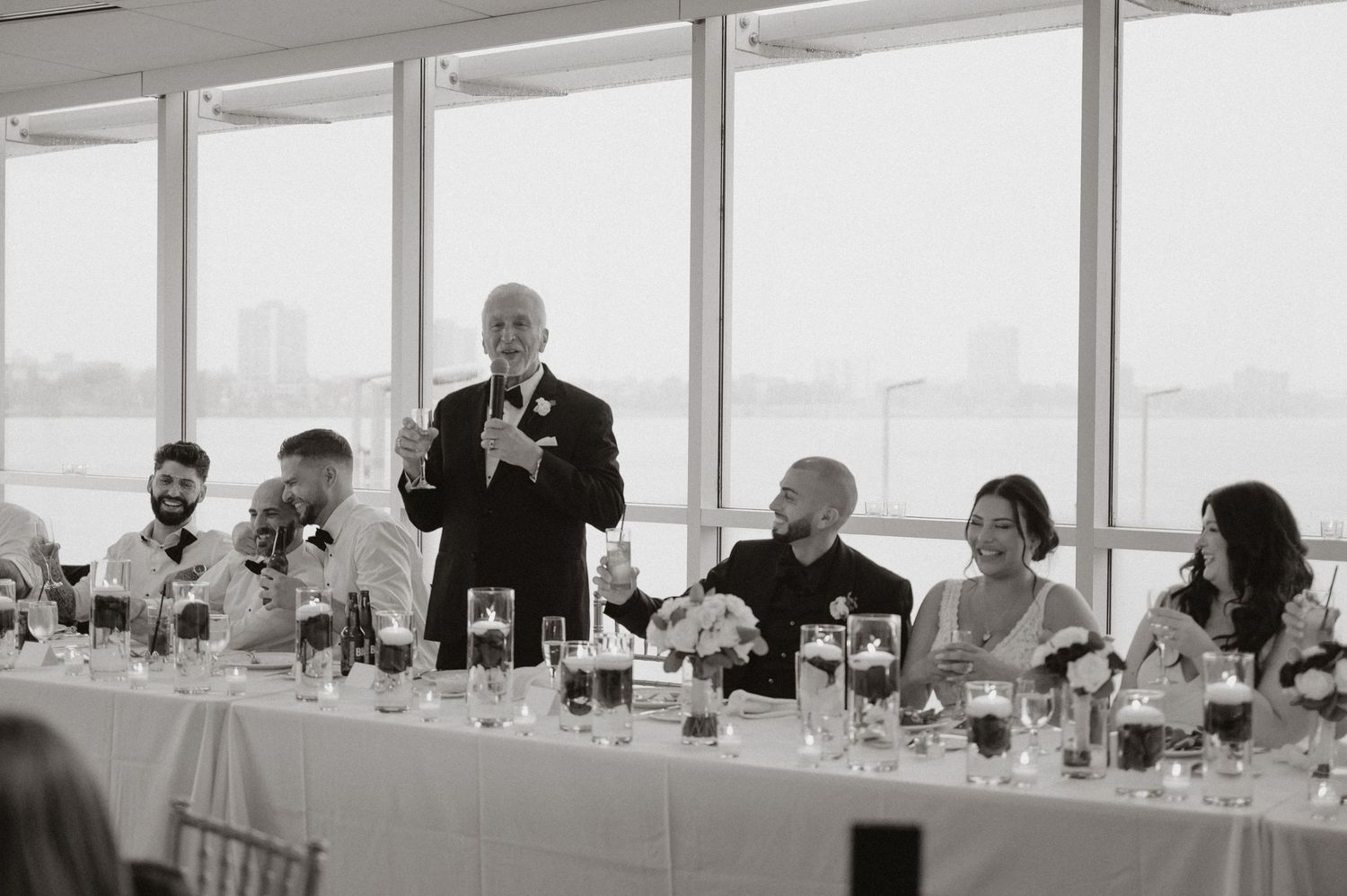 Black and white photo of wedding reception speeches at head table with guests listening intently.