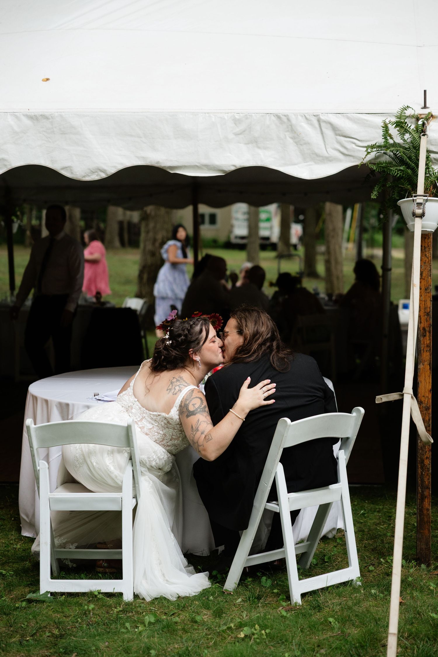 A couple shares a romantic moment under a white tent with folding chairs at an outdoor reception.