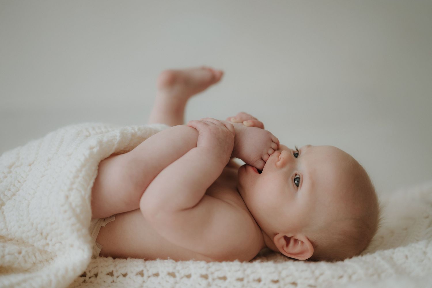Adorable newborn baby lying on a soft white knit blanket, sucking fingers with legs raised in a cozy studio setting.