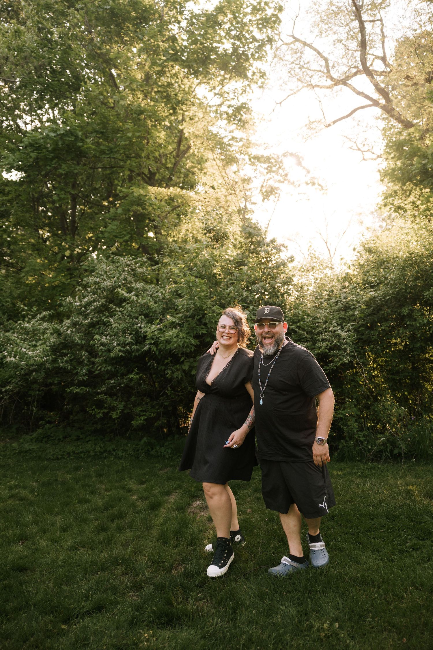 Smiling couple in black outfits pose together in a lush green wooded park setting.