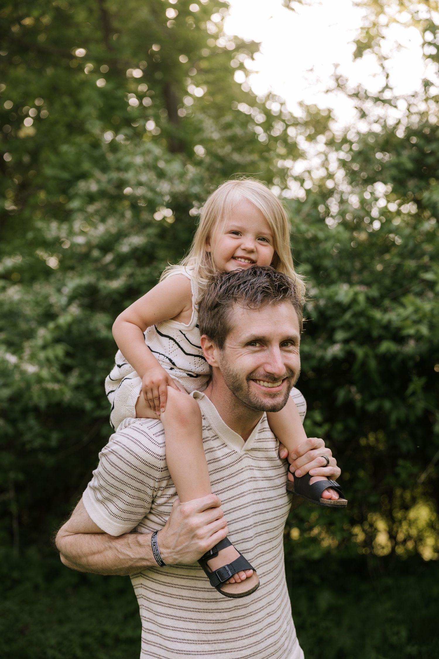 Smiling father carrying laughing toddler daughter on shoulders in sunny green backyard.