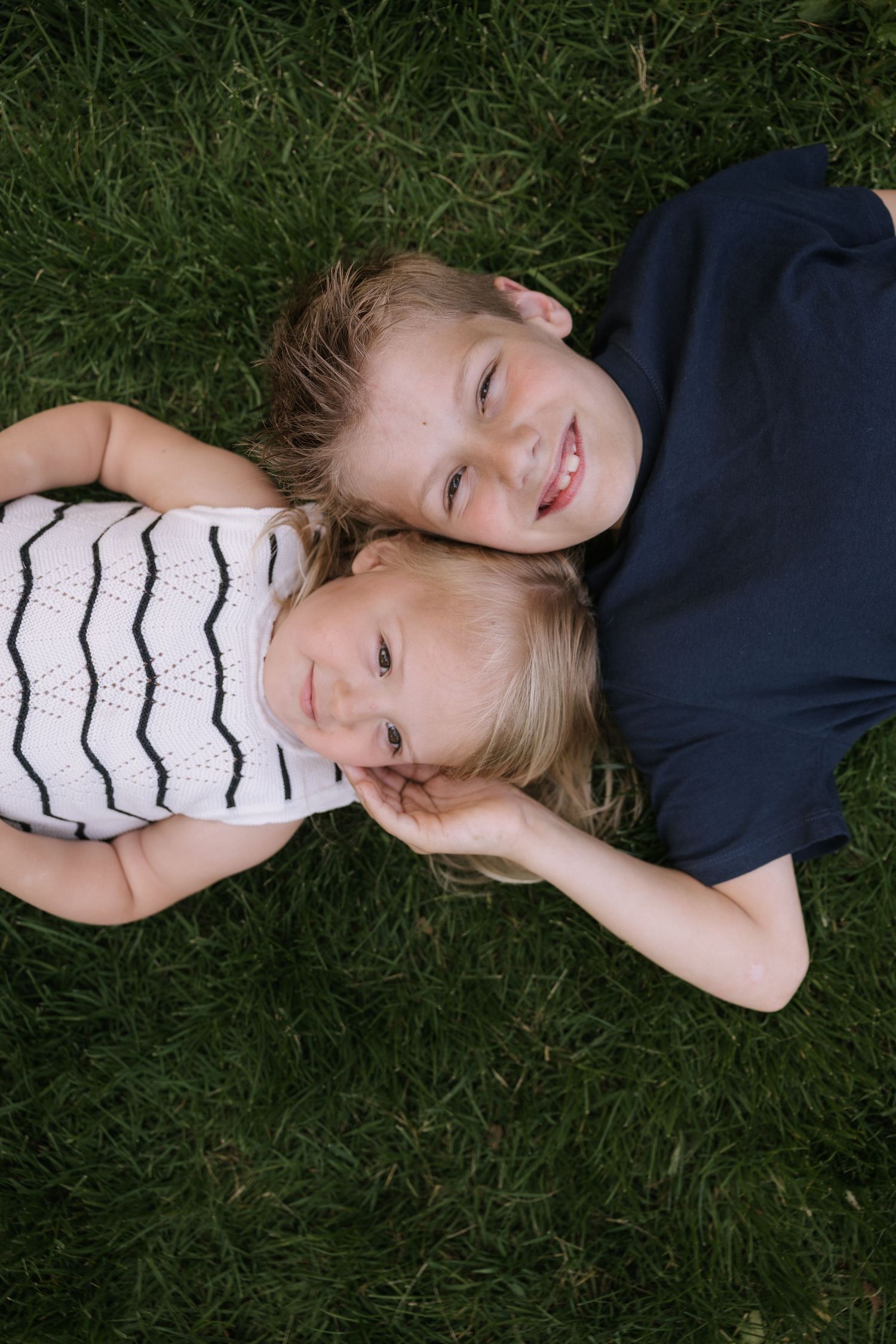 Two smiling blonde children lying head-to-head on green grass looking up at camera.