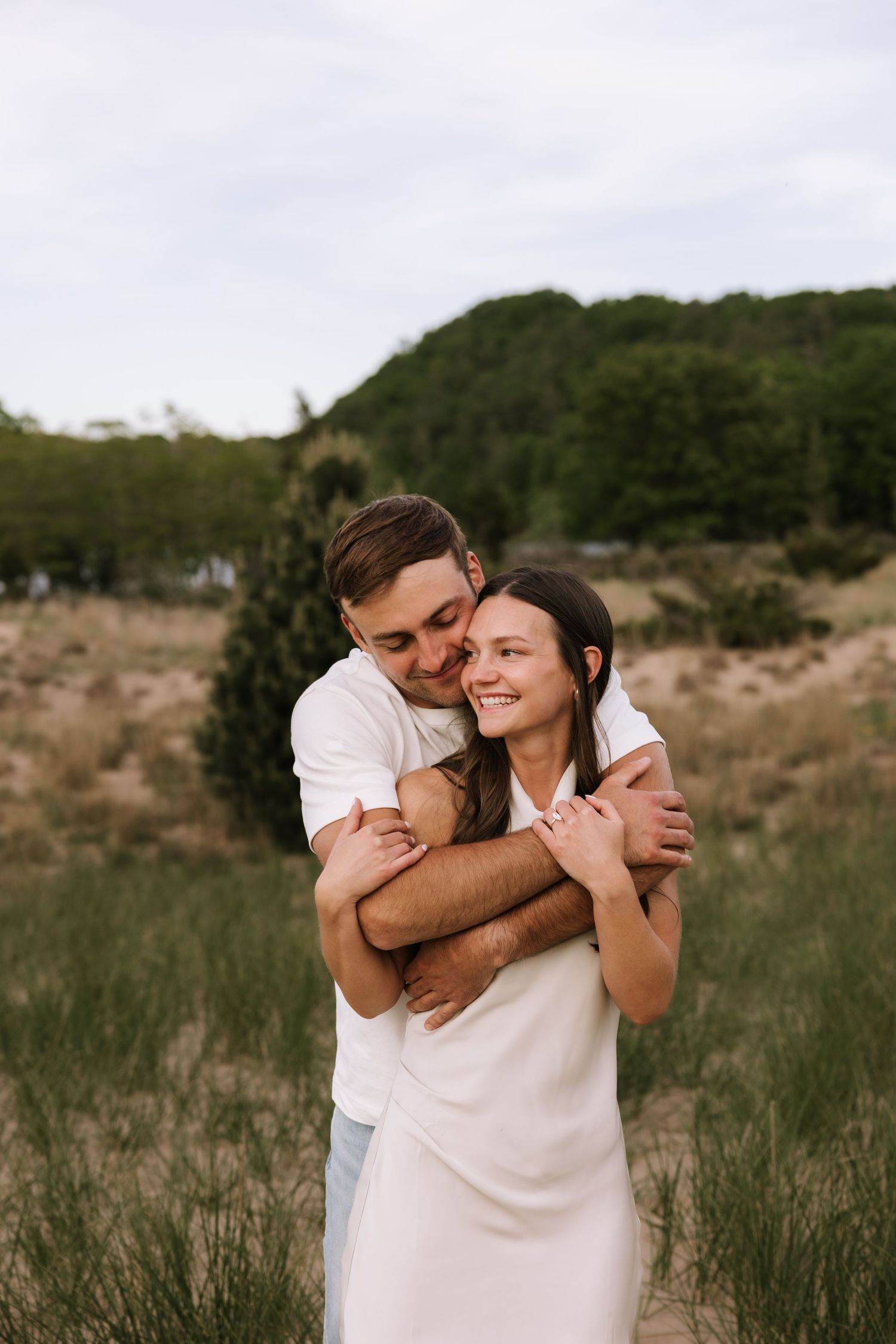 Man hugging woman from behind in grassy field, both smiling, wearing white outfits during outdoor photo session.