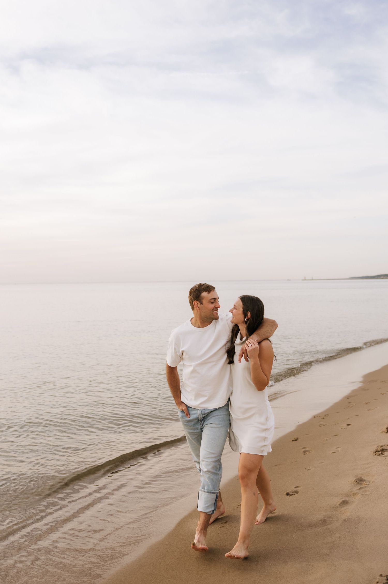 Couple walking barefoot along a calm shoreline, embracing and gazing at each other at sunset.