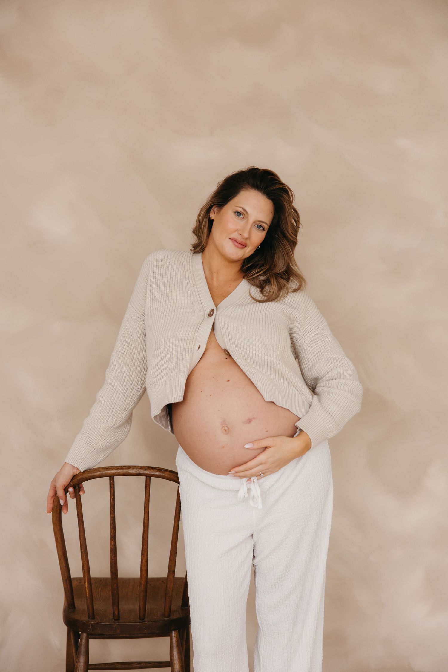 Pregnant woman in beige knit cardigan standing beside wooden chair against textured backdrop.