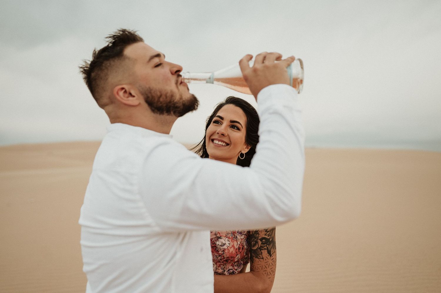 A romantic couple in white and floral outfits share a playful moment together in the desert sand dunes.