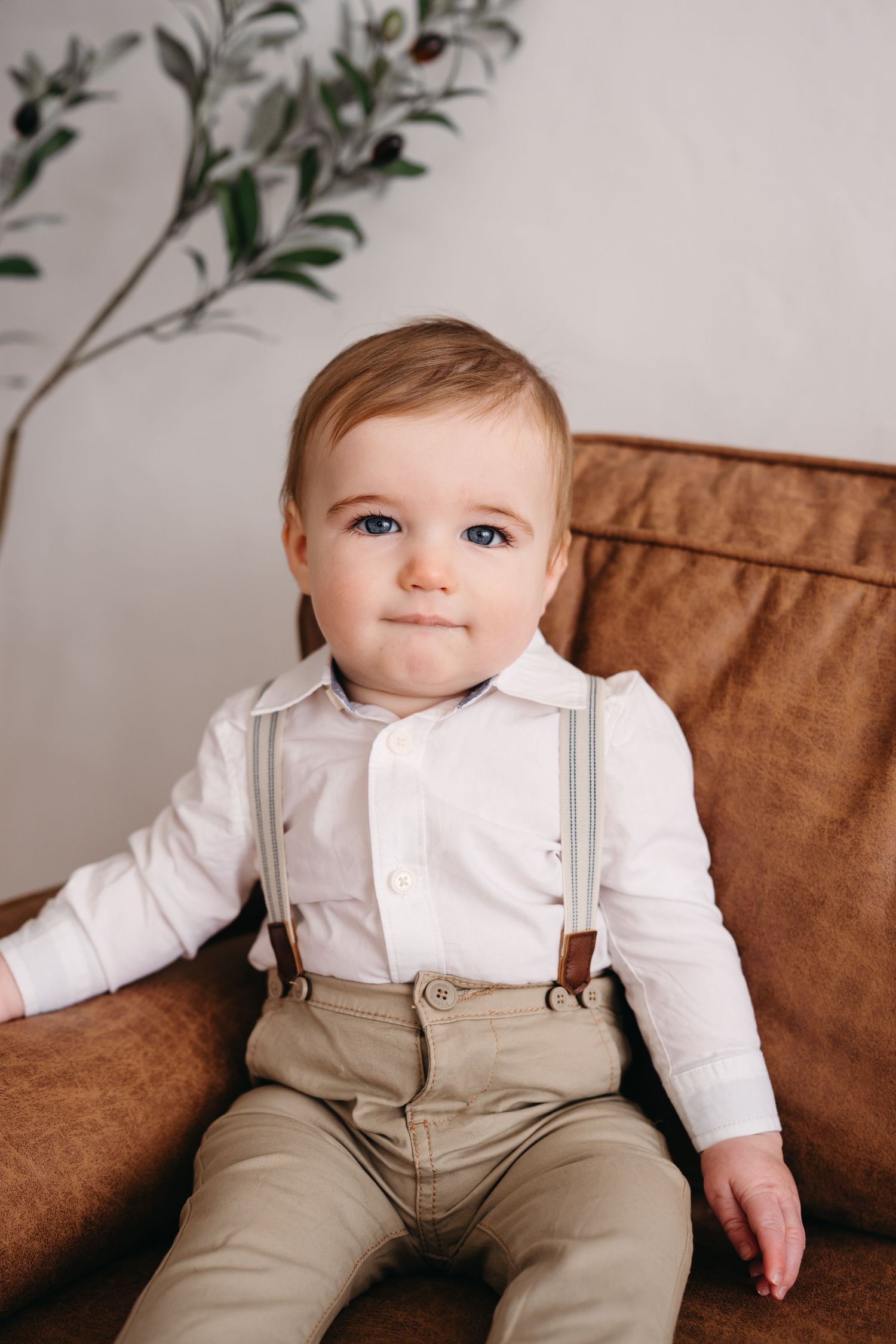 A small child in a white shirt and suspenders sits on a brown leather chair with greenery in the background.