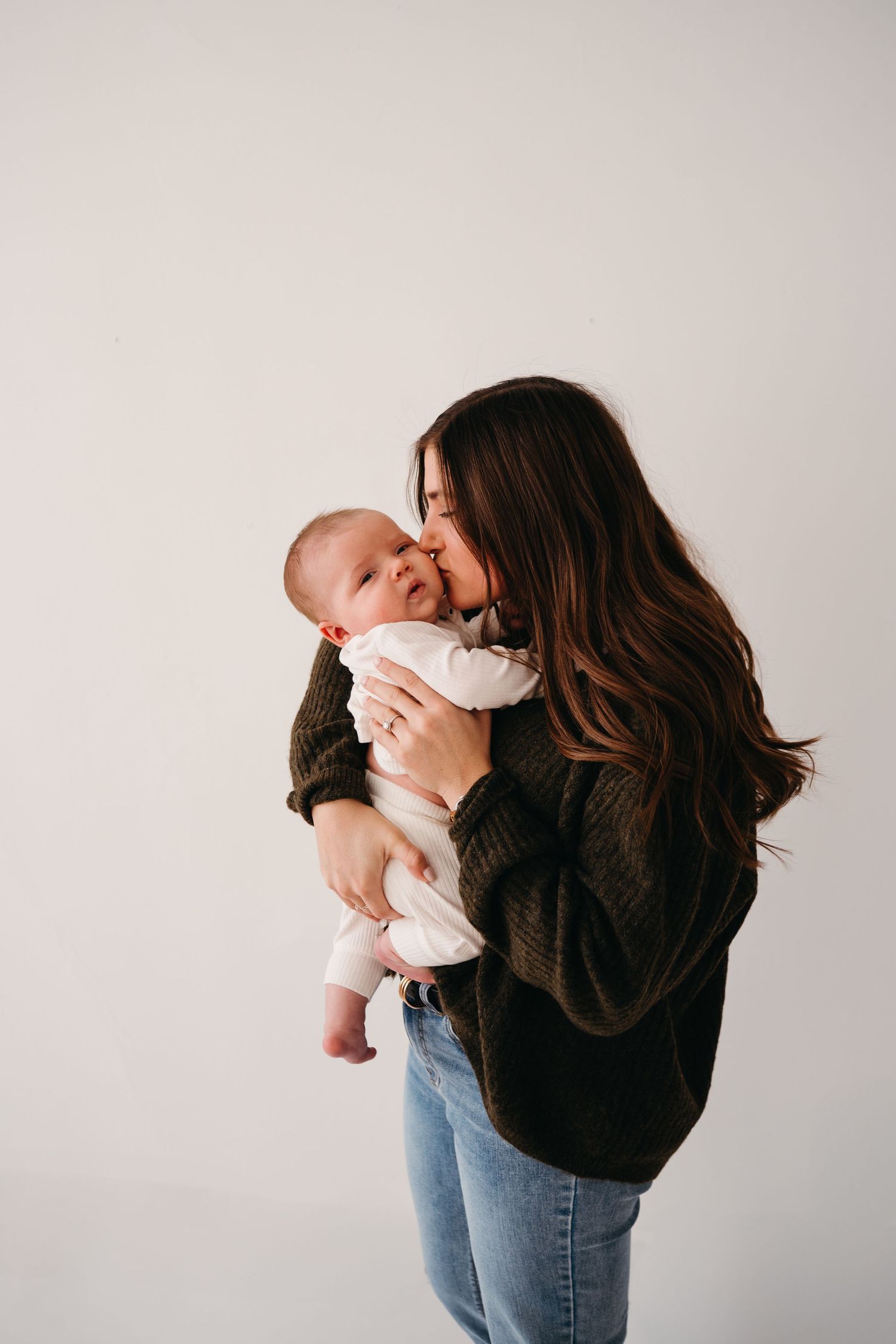 A tender moment captured as someone in a dark sweater cuddles a smiling baby against a white background.