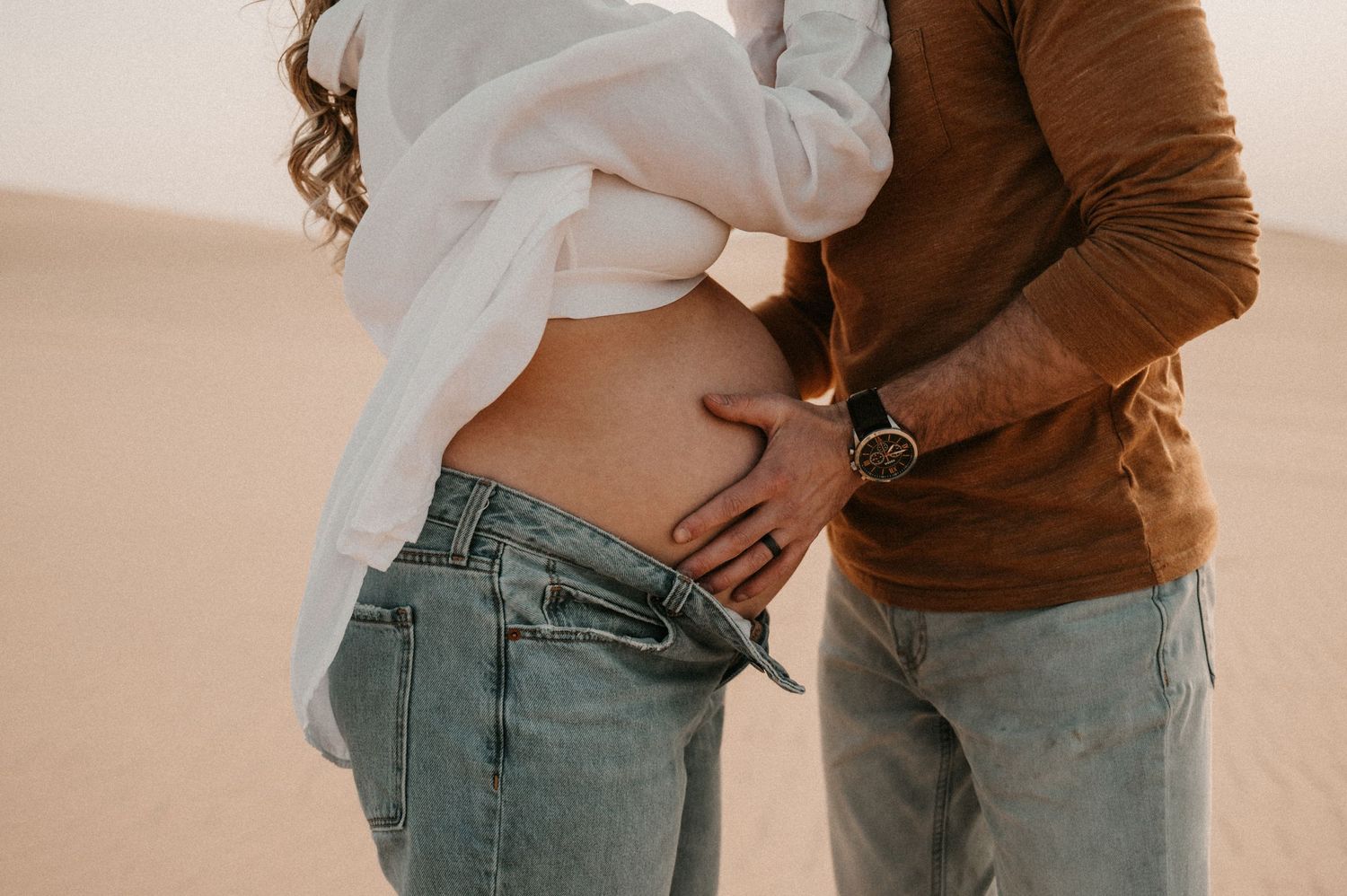 Intimate maternity moment captured between couple on desert sand dunes.
