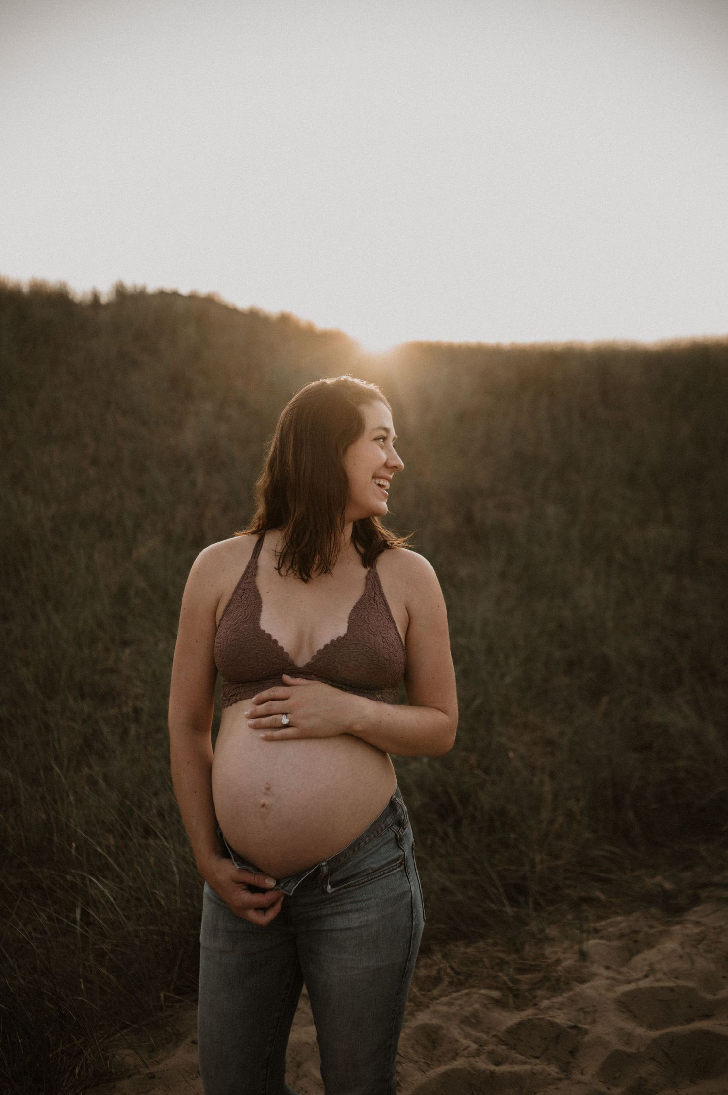 Pregnant woman in brown top and jeans smiles while cradling baby bump at golden hour.