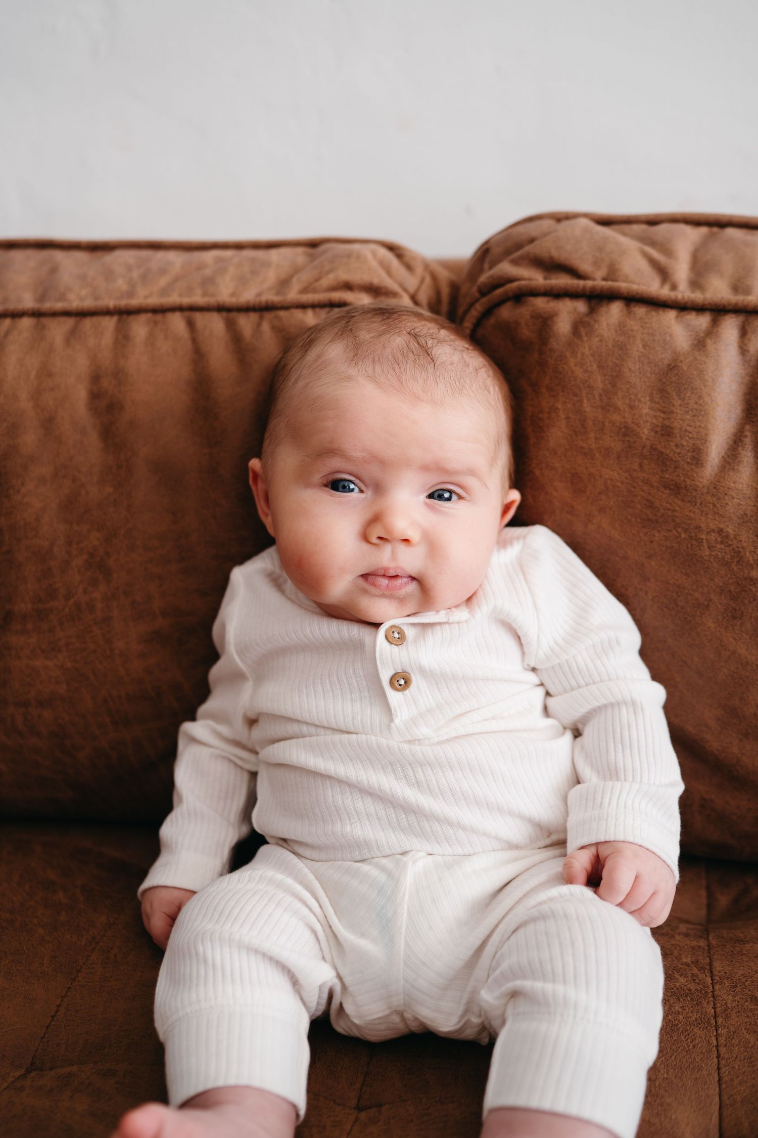 A baby in white ribbed clothing sits on a brown leather couch cushion.