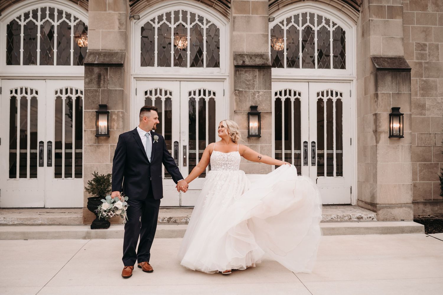 A couple poses playfully outside a grand stone building with elegant arched windows.