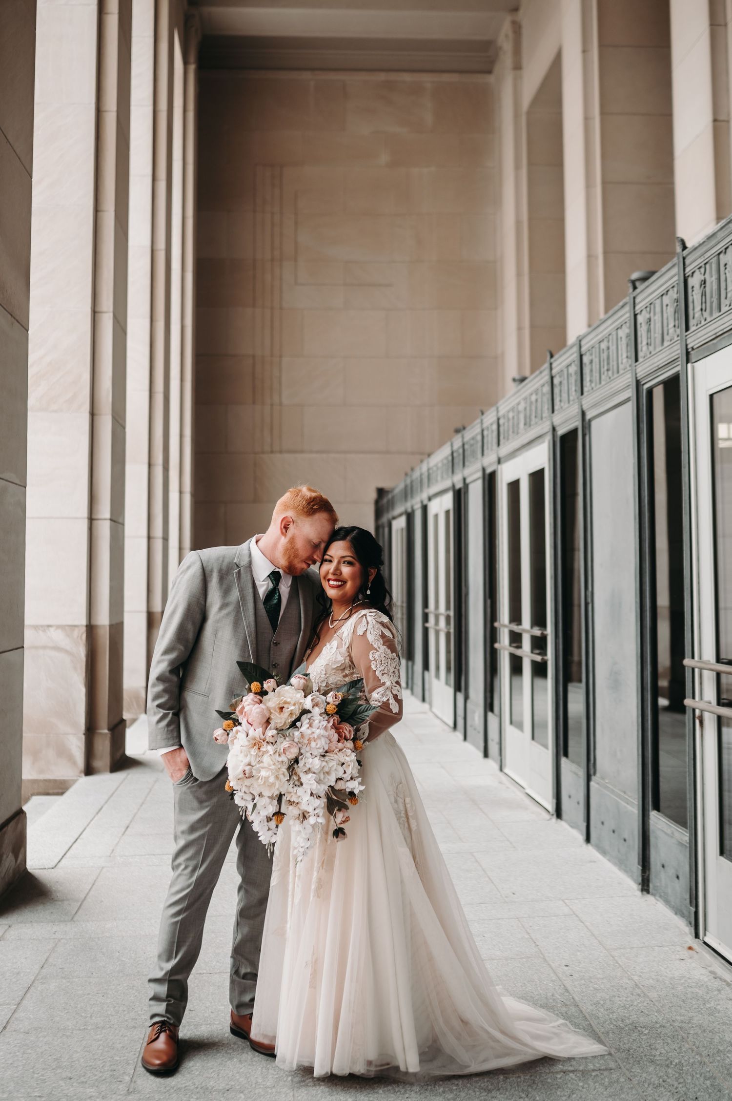 Newlyweds share intimate moment in elegant hallway with stone columns and glass doors.