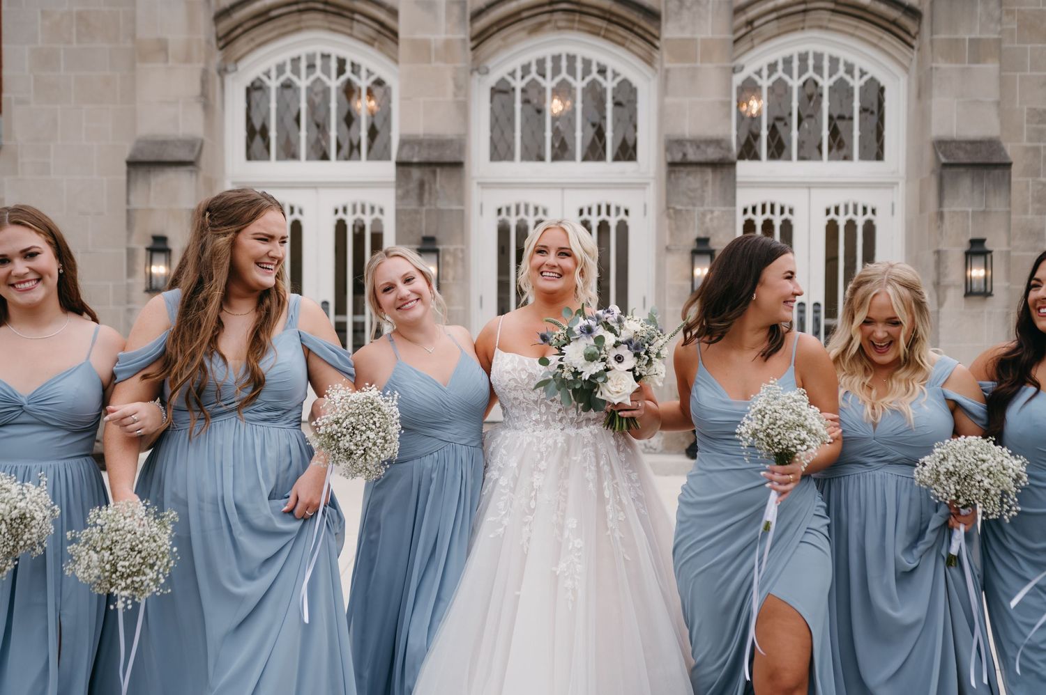 Bridal party laughs together wearing matching dusty blue gowns with white floral bouquets.