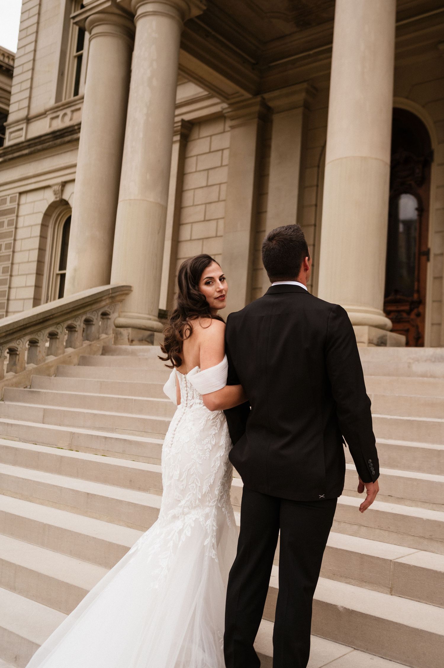 A couple in wedding attire walks up grand stone steps of a historic building.