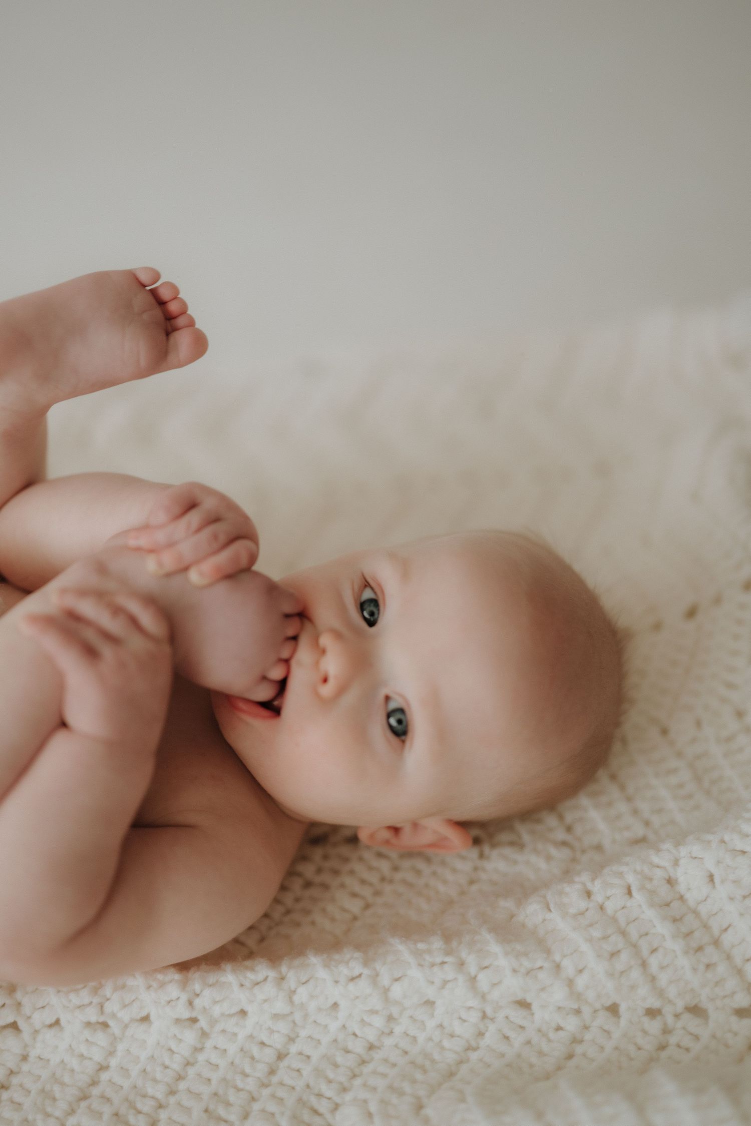Blue-eyed baby lying on cream crochet blanket, chewing on toes playfully.