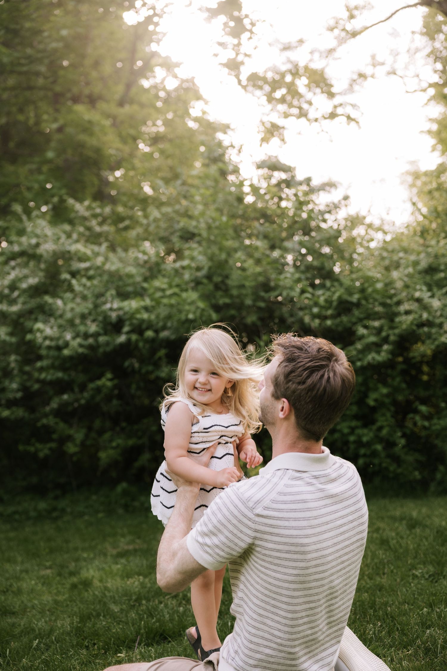 Father sitting on grass lifting laughing blonde toddler girl in striped dress outdoors.