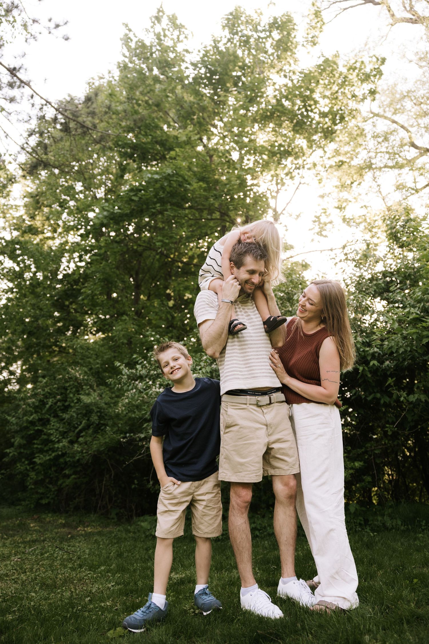 Happy family of four posing outdoors with dad giving child a shoulder ride in green backyard.