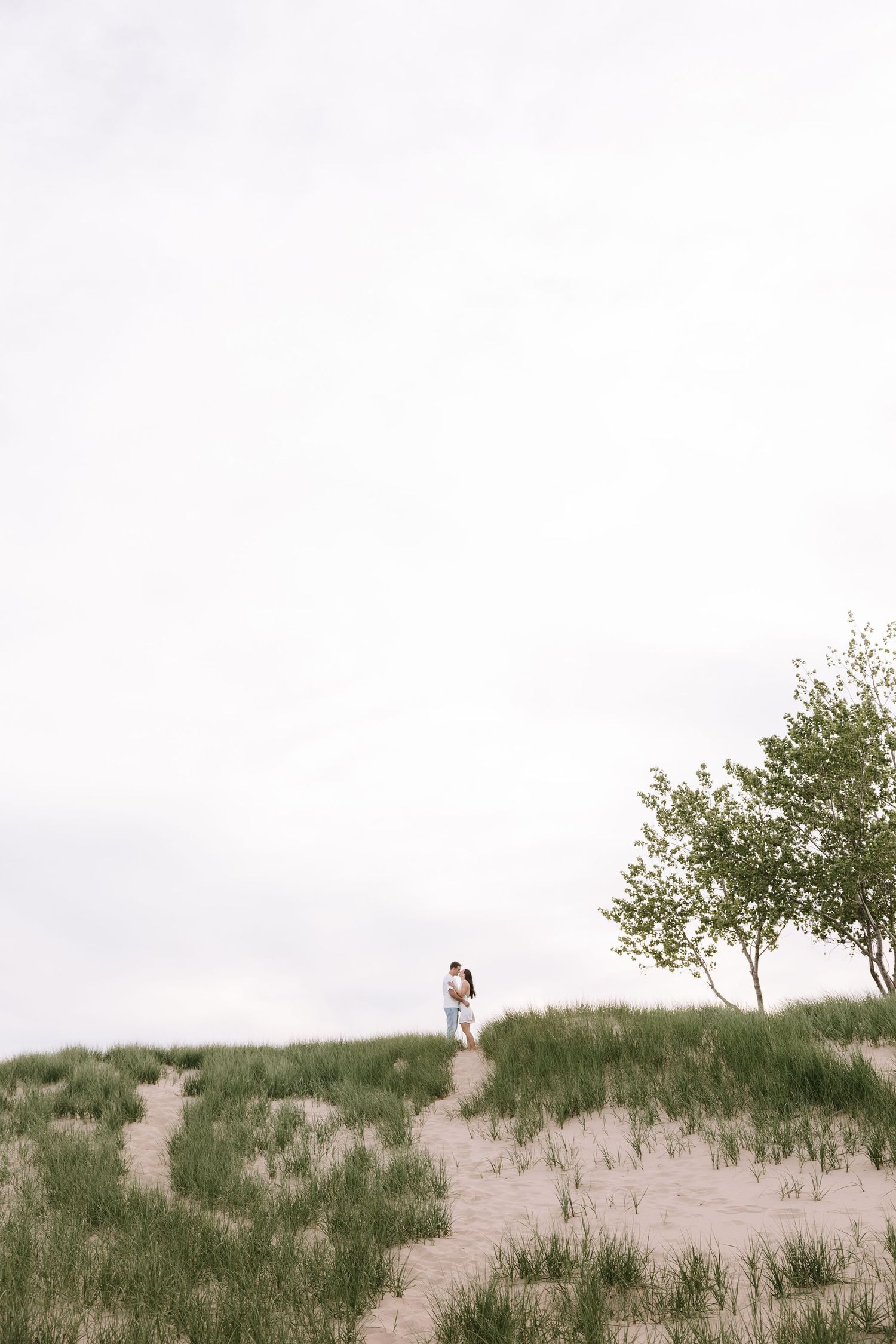 Couple kissing on grassy sand dune path, small tree nearby, vast overcast sky above scenic landscape.
