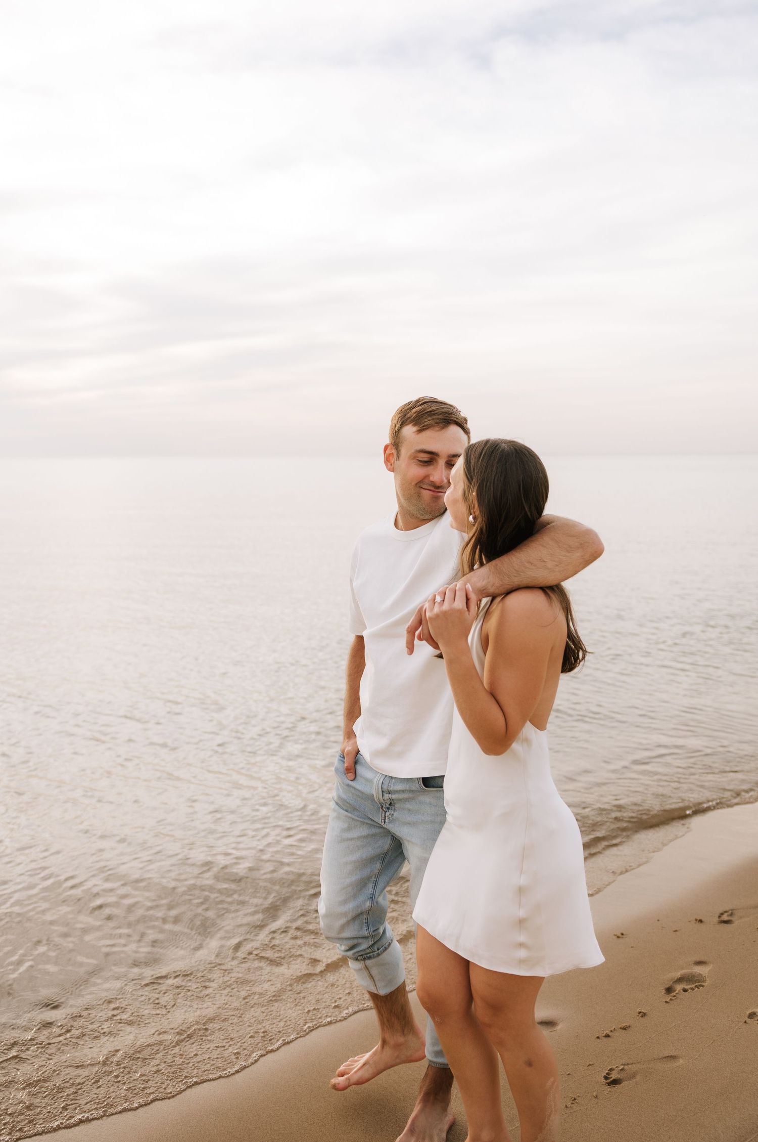 A couple in white outfits embracing and smiling on a sandy beach at sunset near calm ocean waters.