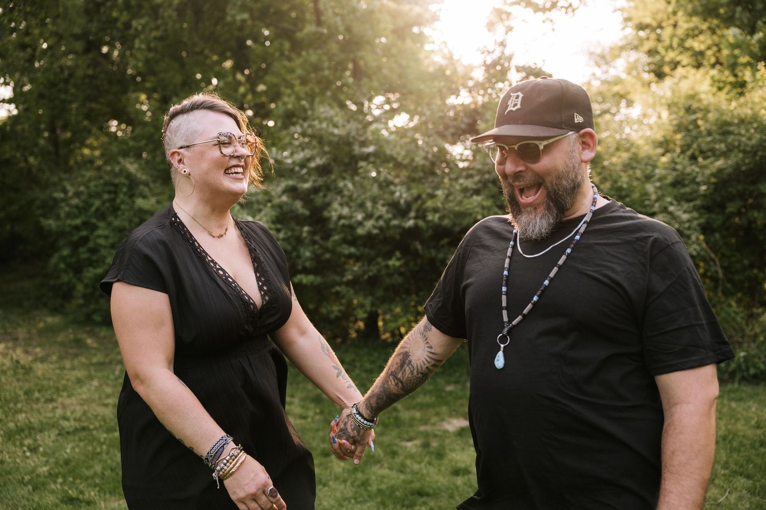 Tattooed couple holding hands and laughing in sunny outdoor garden.