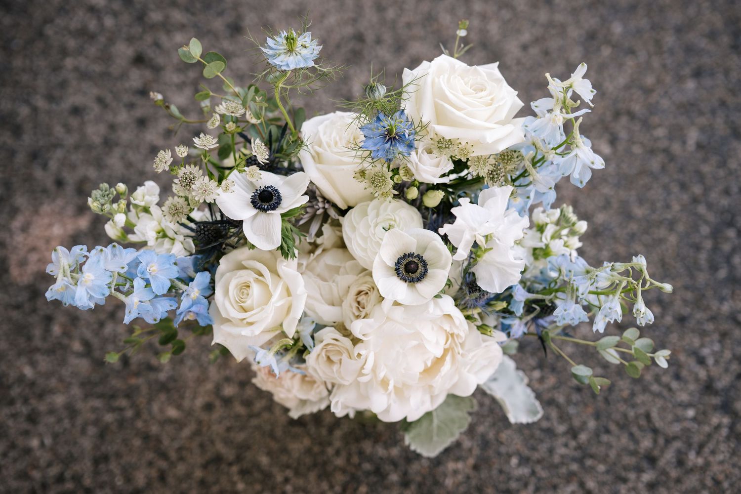 Delicate floral wedding bouquet arrangement with white roses and small blue forget-me-not flowers on a textured grey surface.